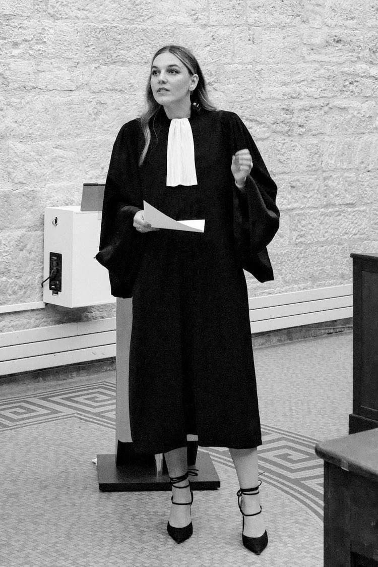 Femme en robe d'avocat parlant lors d'un procès ou d'une audience, tenant un document, dans une pièce aux murs en pierre.