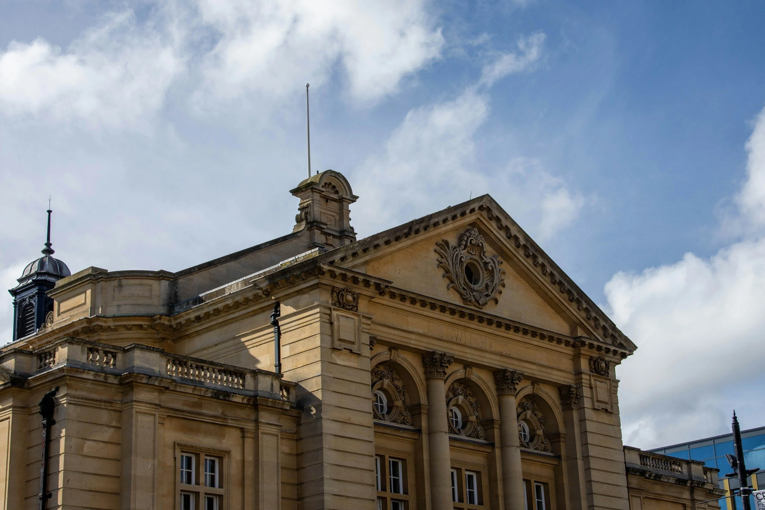 Façade d'un ancien bâtiment en pierre avec des colonnes et un fronton orné, sous un ciel partiellement nuageux.