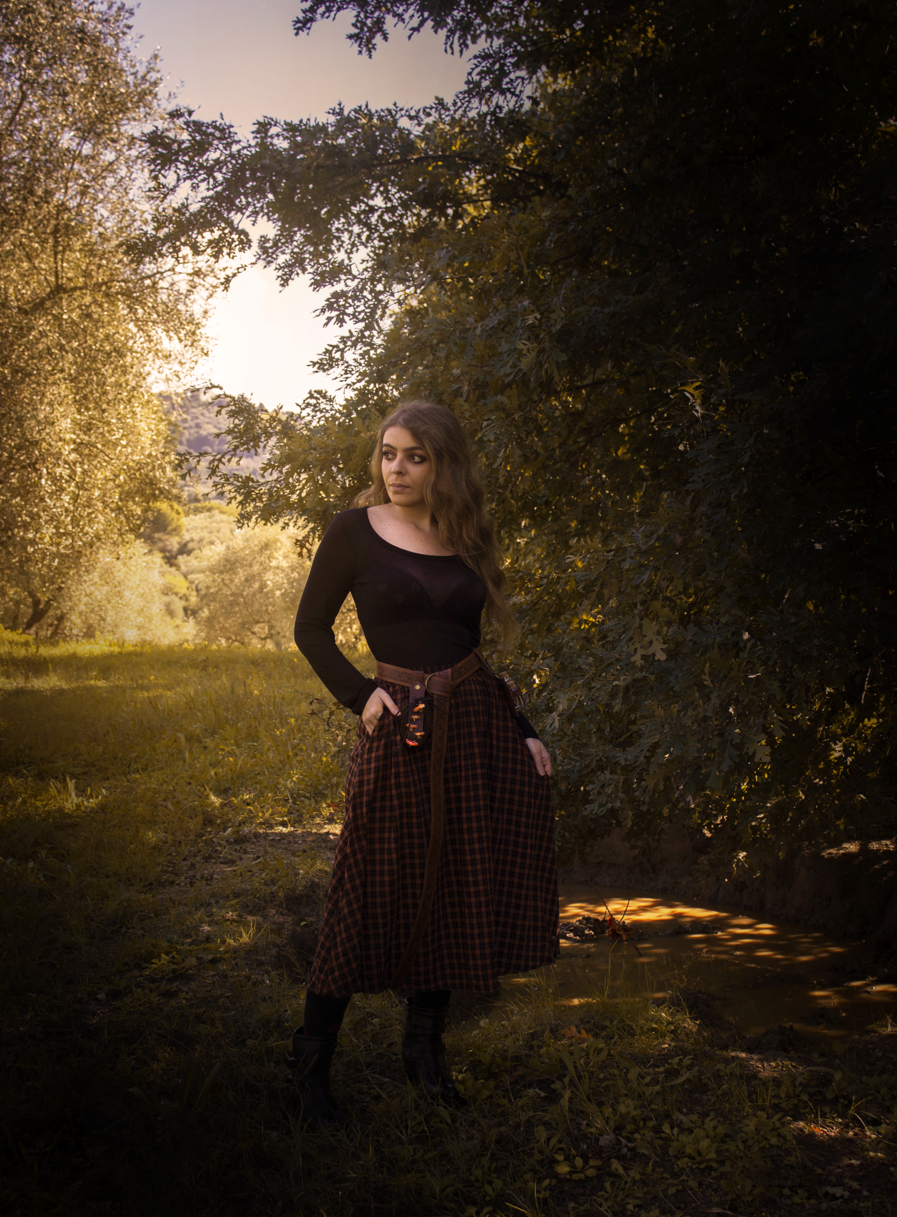 A young woman with long wavy hair standing outdoors in a wooded area during sunset, wearing a black top and a long plaid skirt.