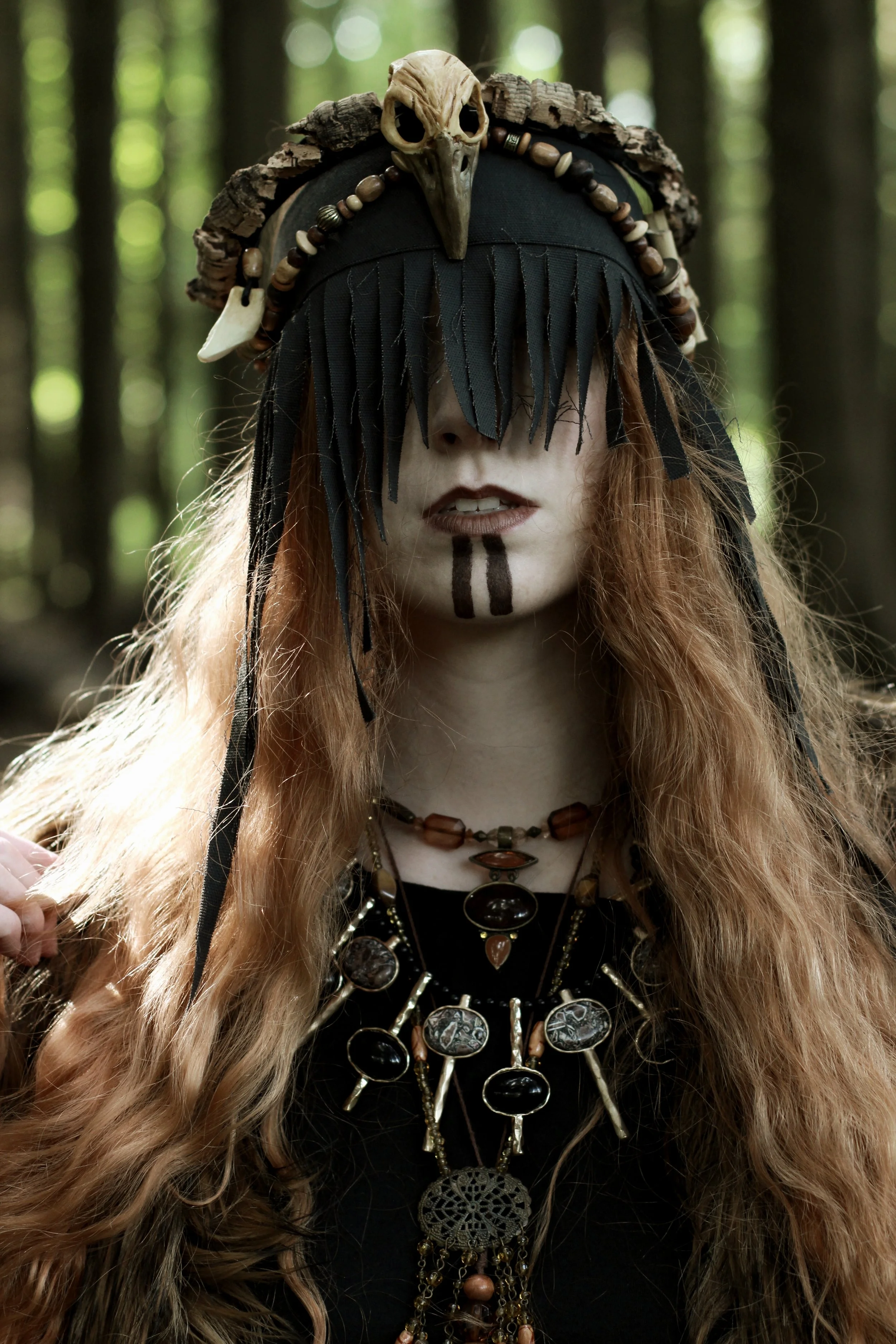 Donna con trucco bianco e linee nere sul viso, indossa un copricapo decorato con ossa e un cranio, con capelli lunghi e ricci, indossa diversi gioielli e abiti neri, ambientata in un bosco.