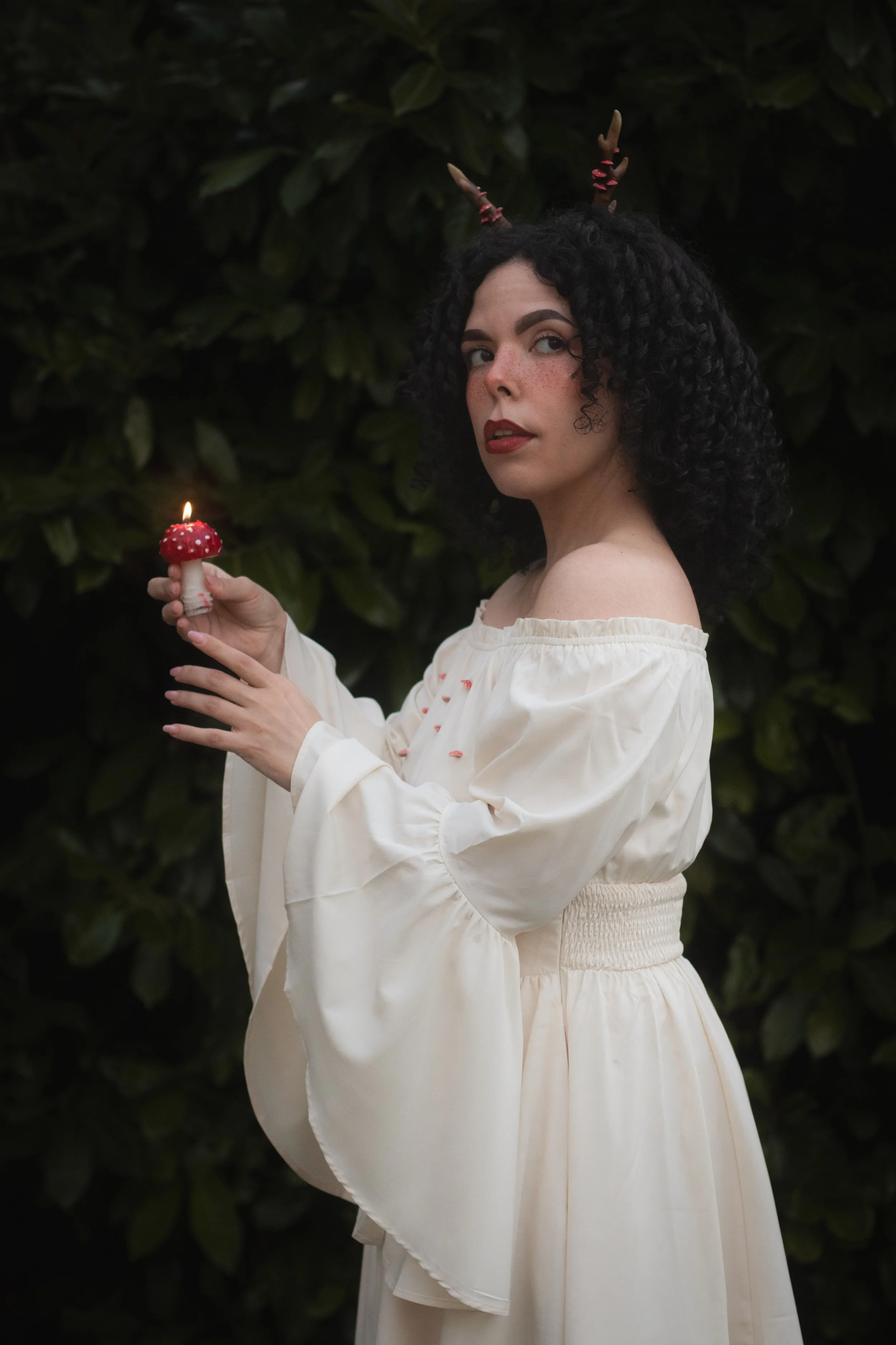 Donna con capelli ricci neri e pelle chiara, vestita in abbigliamento bianco con maniche a campana, tiene una piccola lampada a forma di fungo rosso con luci, in piedi davanti a un cespuglio.