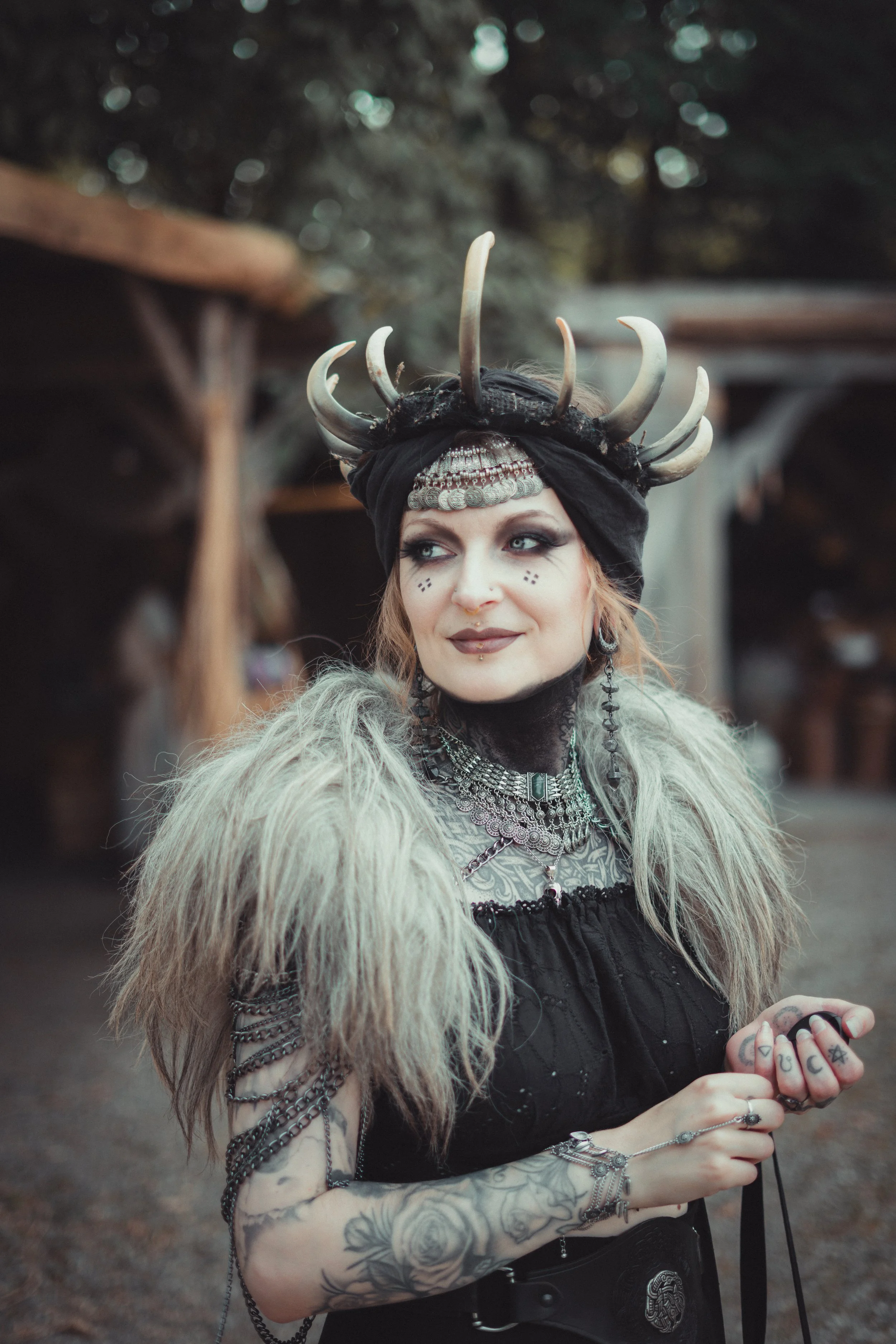 Una donna con trucco e tatuaggi indossa un costume con un copricapo decorato con corna e monete, e accessori metallici, sorride e dà un'occhiata di lato.