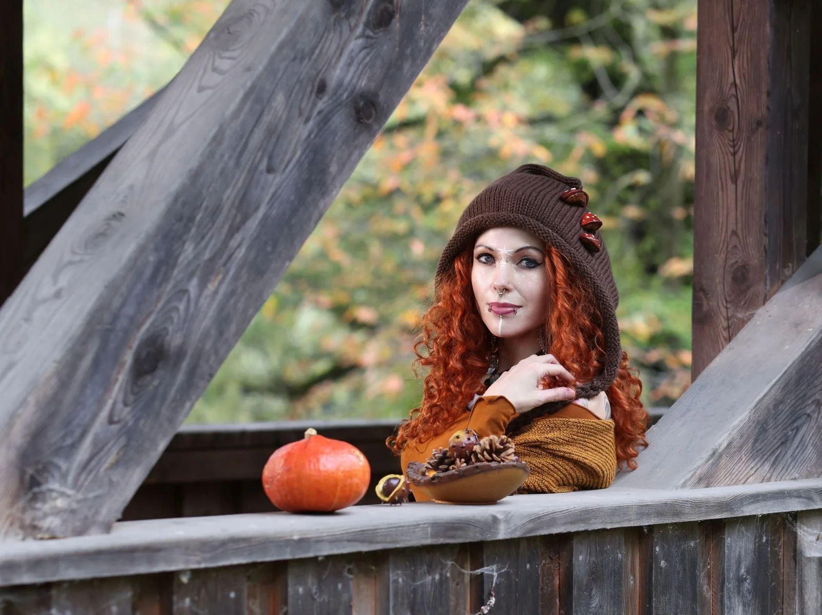 Donna con capelli ricci rossi seduta su una struttura di legno, indossa un cappello marrone decorato con pietre, trucco artistico, e ha piercing. Davanti a lei, una zucca e una ciotola con pigne e sferette decorative.