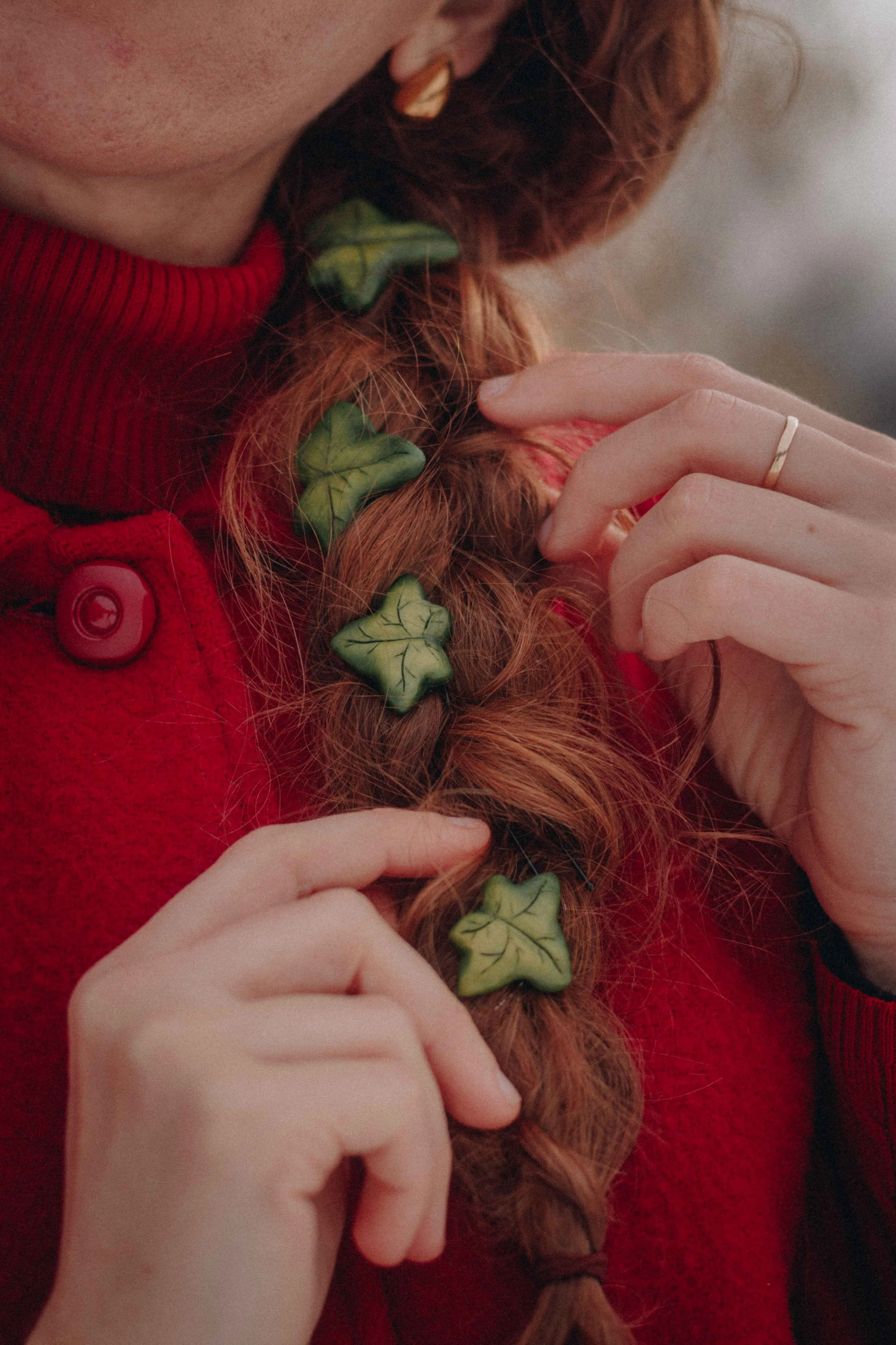 Close-up di una persona con capelli ricci rossi decorati con foglie di edera finte, mentre regge una ciocca di capelli.