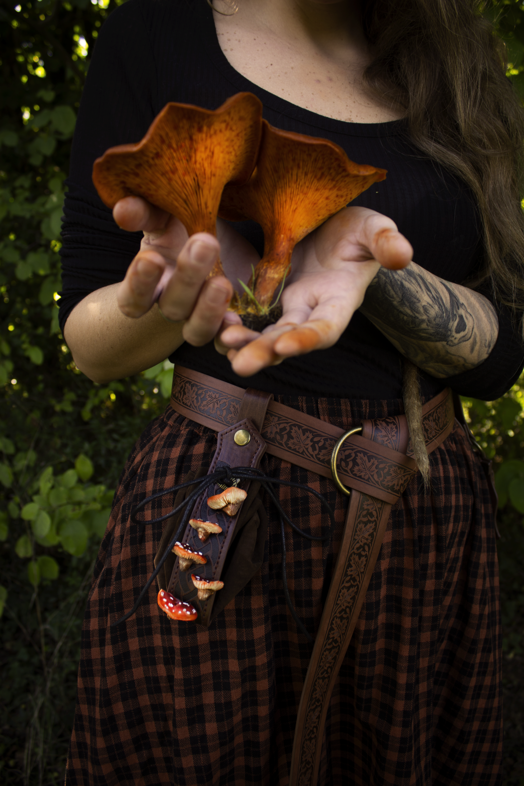Donna con abbigliamento in stile fantasy che tiene in mano due funghi giallo/arancio e marrone, circondata da vegetazione.