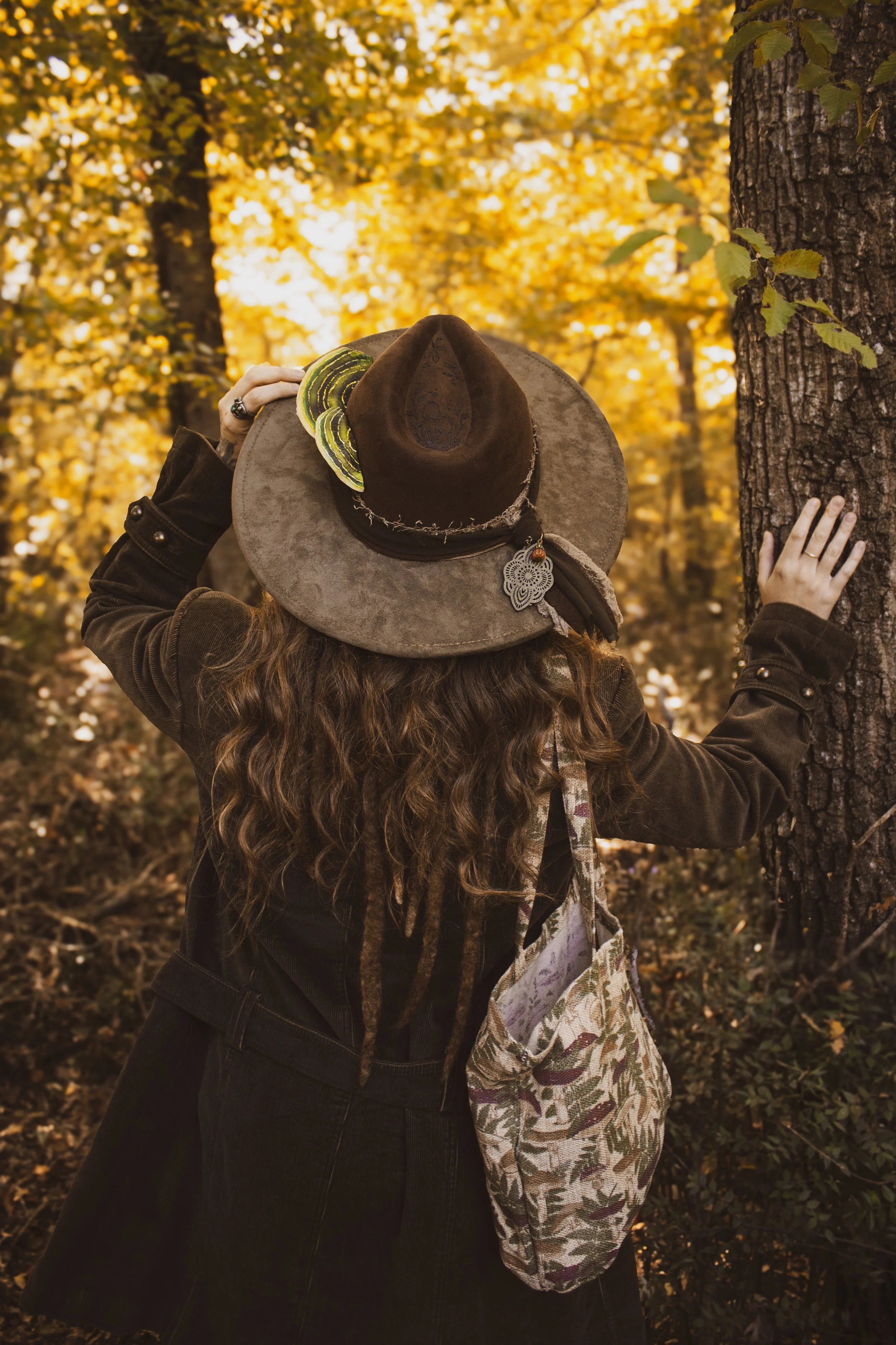 Una donna con capelli ricci e vestiti scuri si trova in un bosco autunnale, appoggiata a un albero, con un cappello di feltro marrone decorato e una borsa di stoffa con disegni floreali.