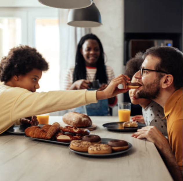 Child feeding a man a cookie during breakfast with family, on a table with donuts, pastries, orange juice, and a child in the background