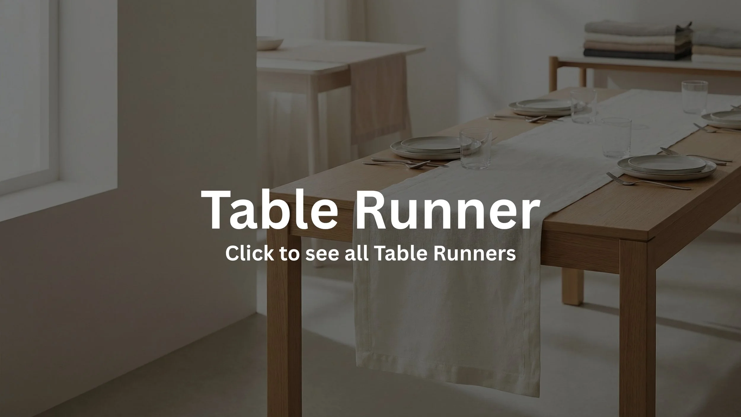 A wooden table with a light-colored table runner, set with plates, knives, forks, and water glasses, in a bright room with natural light.