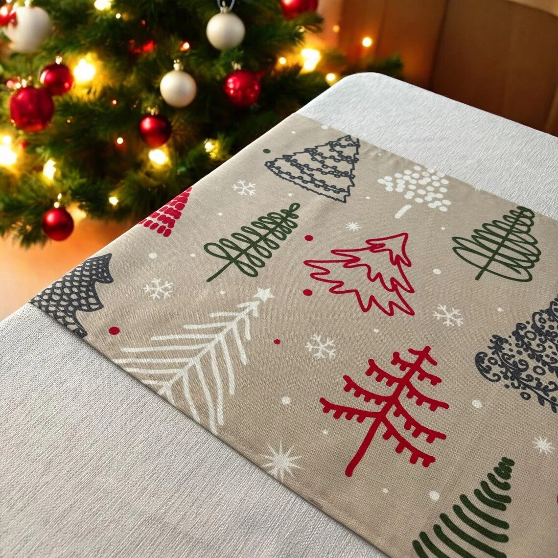 A holiday table setting with a beige napkin featuring Christmas tree and snowflake patterns, placed on a white tablecloth against a backdrop of a decorated Christmas tree with red and white ornaments and lights.