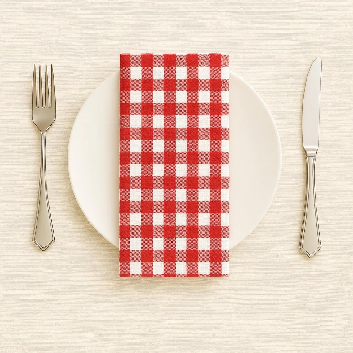 Place setting with a white plate, silver fork and knife, and a red and white checkered paper napkin on a beige tablecloth.