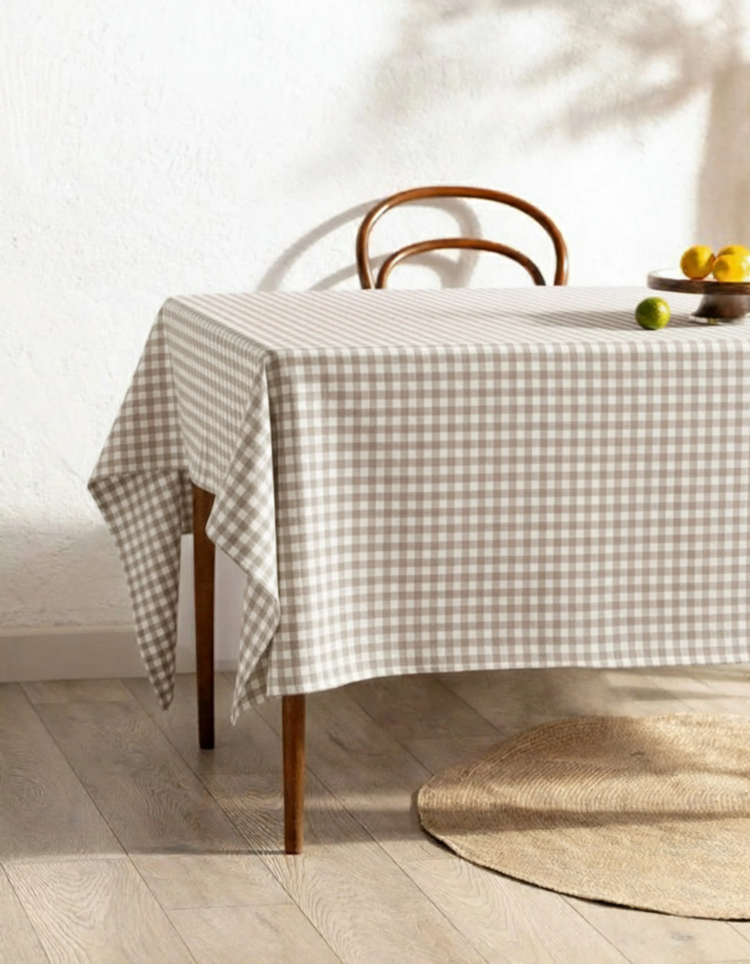 A dining table with a beige checkered tablecloth, brown wooden chairs, and a small plate of lemons and limes on top, with a beige rug underneath.