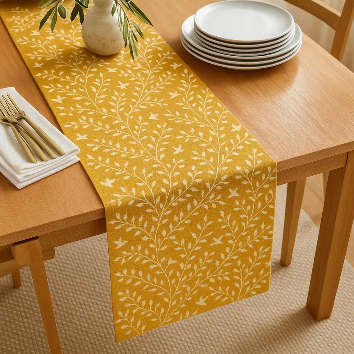 Wooden dining table with a yellow and white leaf-patterned table runner, a white vase with green leaves, a stack of white plates, and gold cutlery on a white napkin.