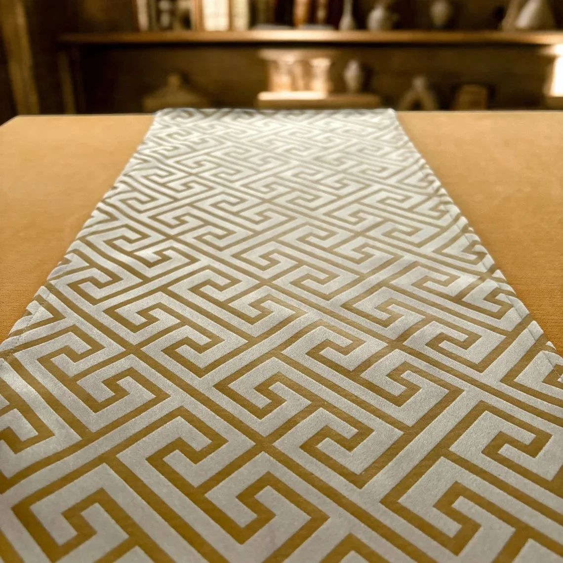 A decorative table runner with a gold and white geometric Greek key pattern on a wooden table.