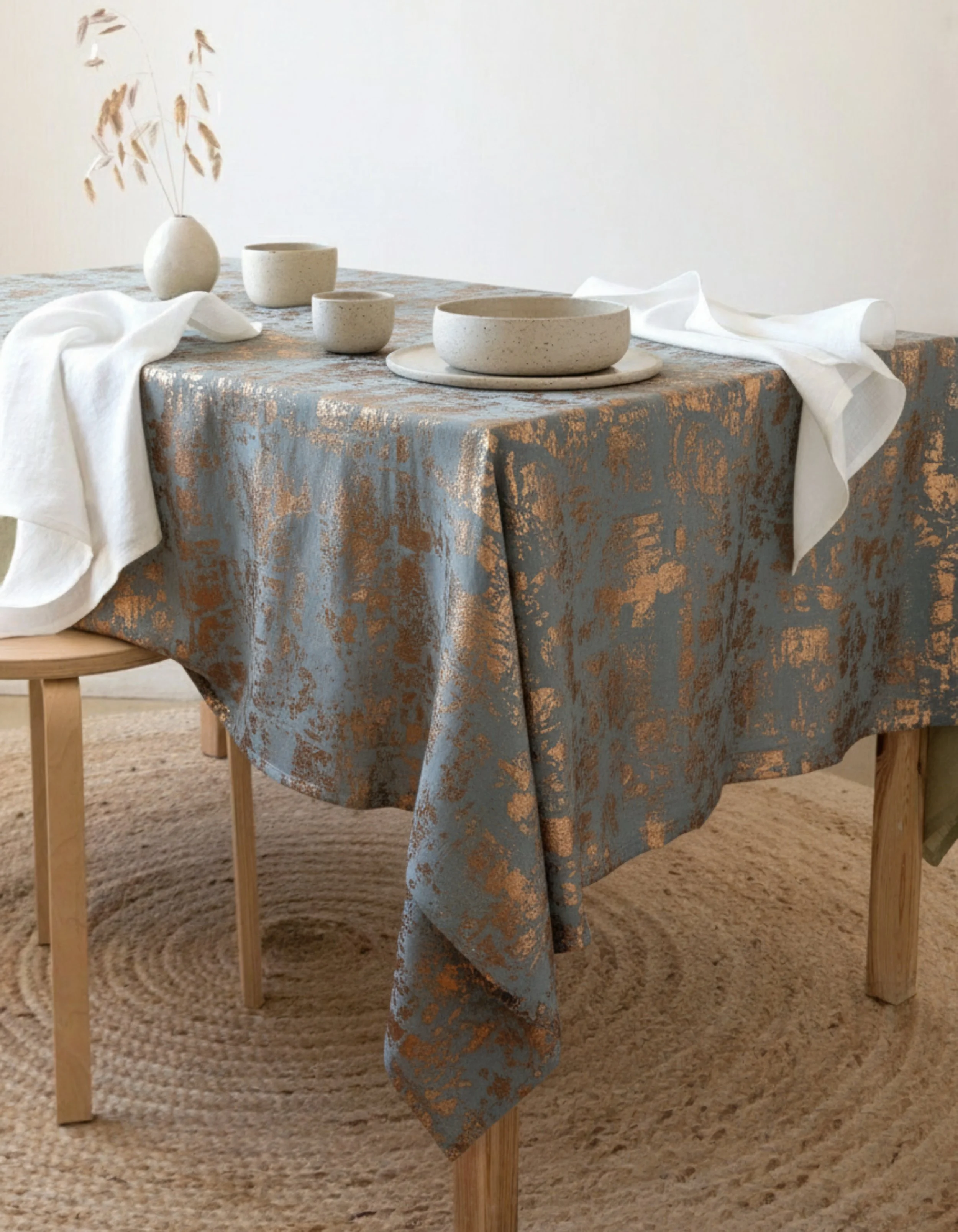 A dining table with a patterned tablecloth, set with ceramic bowls, cups, and a plate, alongside a white cloth, with a vase of dried plants on the table, and a wooden chair partially visible.