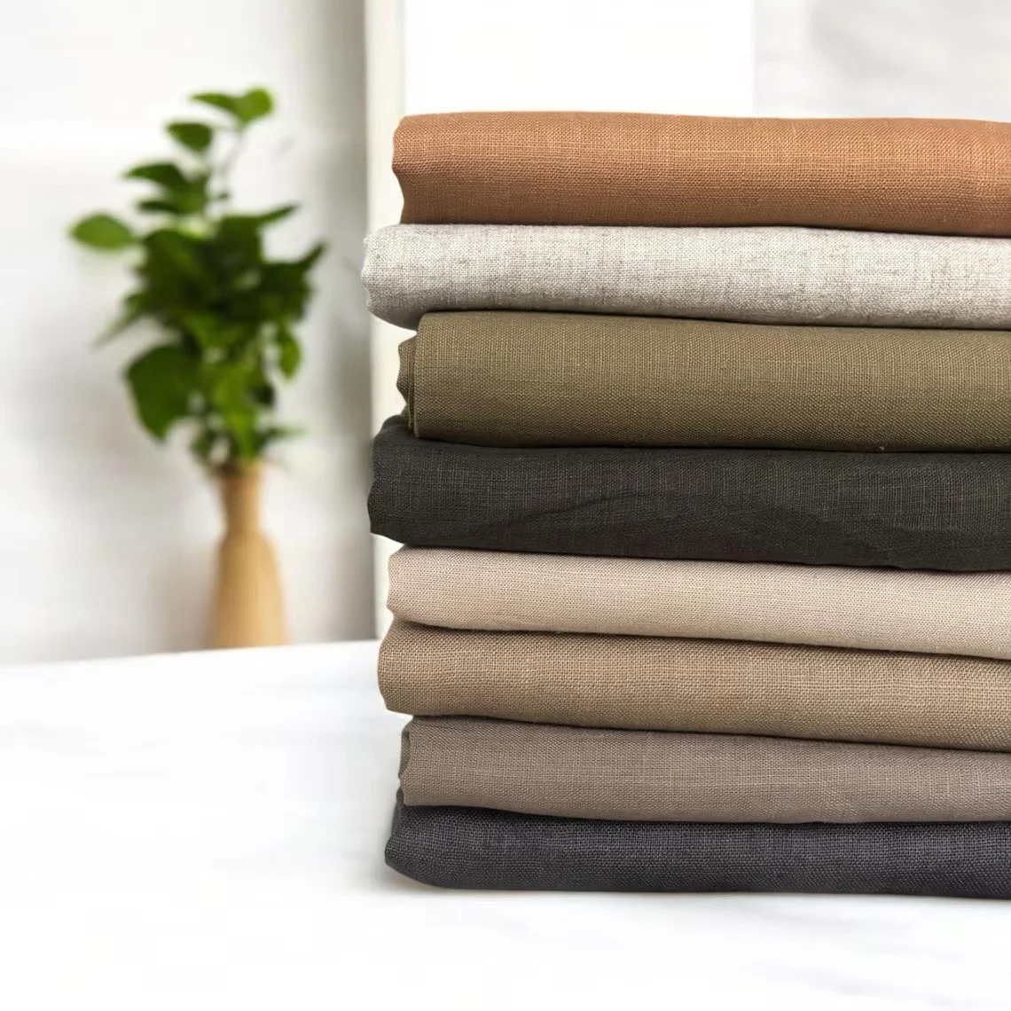 Stack of folded neutral-colored fabrics or textiles with a potted plant in the background.