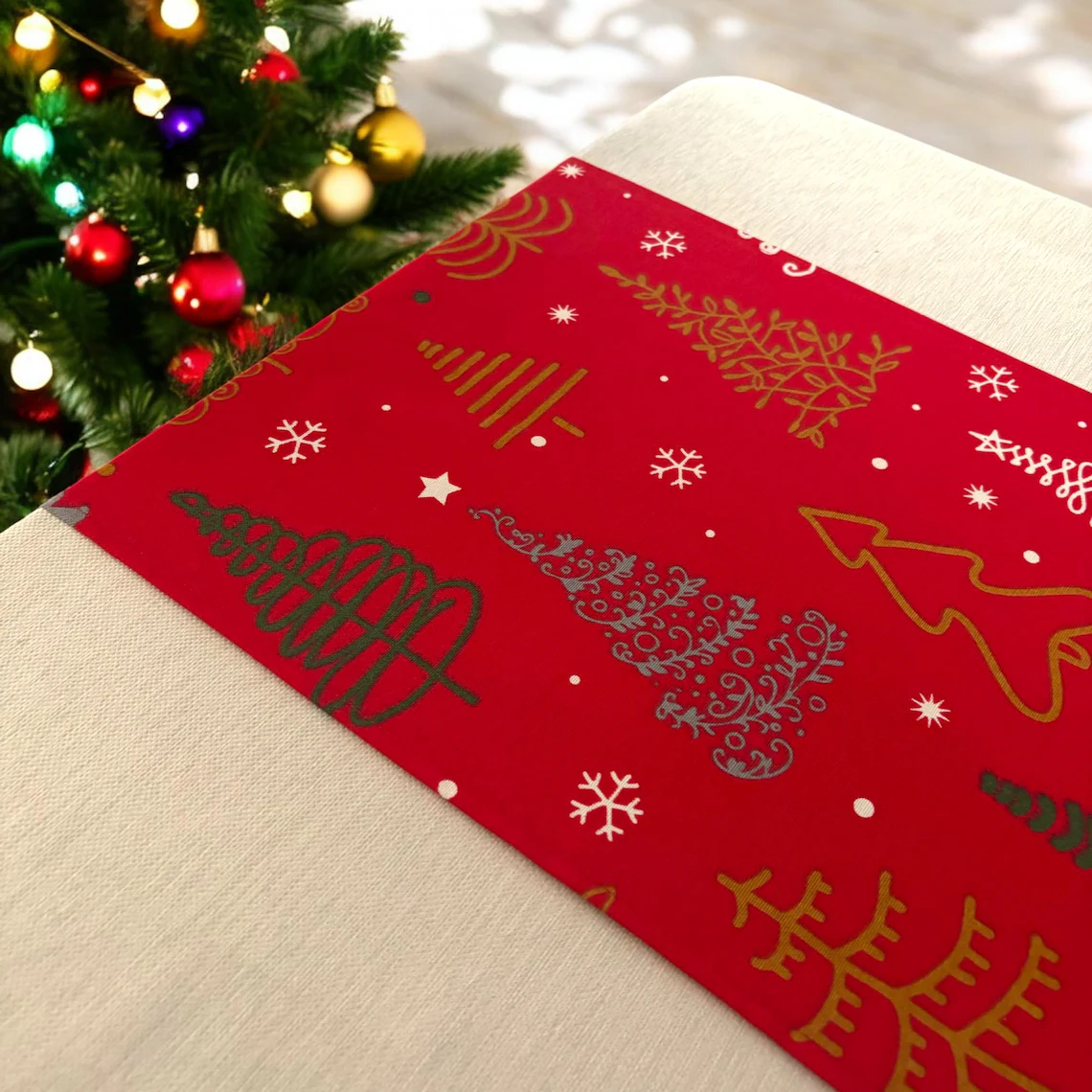 Close-up of a festive red napkin with holiday-themed gold and white drawings, placed on a tablecloth near a decorated Christmas tree with red, gold, and purple ornaments.