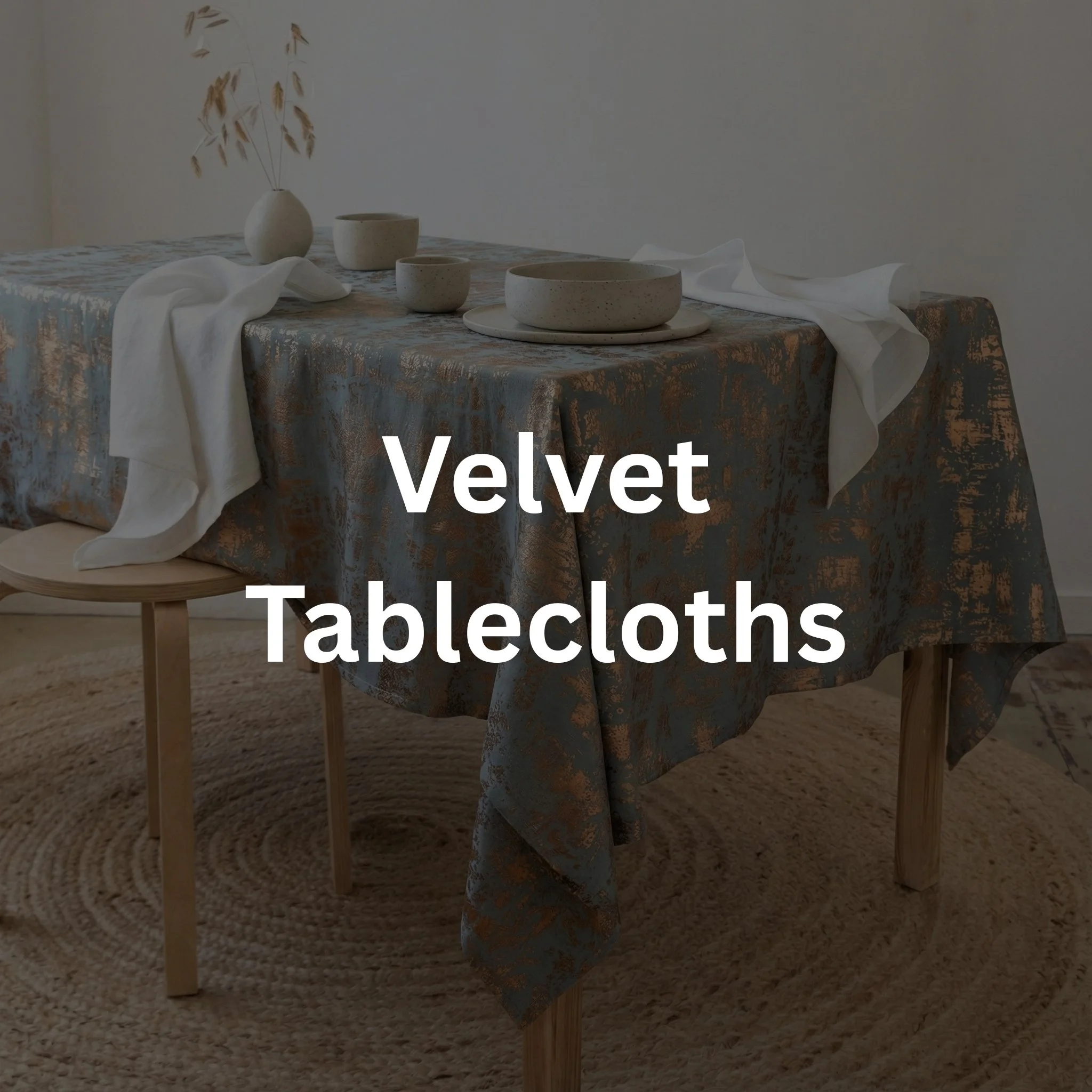 A dining table with a velvet tablecloth, ceramic dishes, and a white cloth, surrounded by wooden chairs, with a neutral wall and a vase with dried plants in the background.