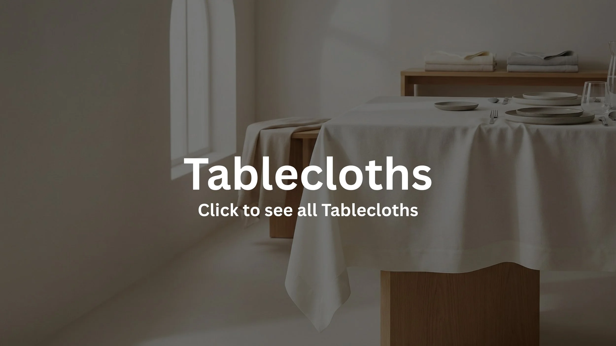 A dining table covered with a white tablecloth, set with plates, forks, knives, spoons, and glasses, in a minimalist, softly lit room with natural light coming through a window.