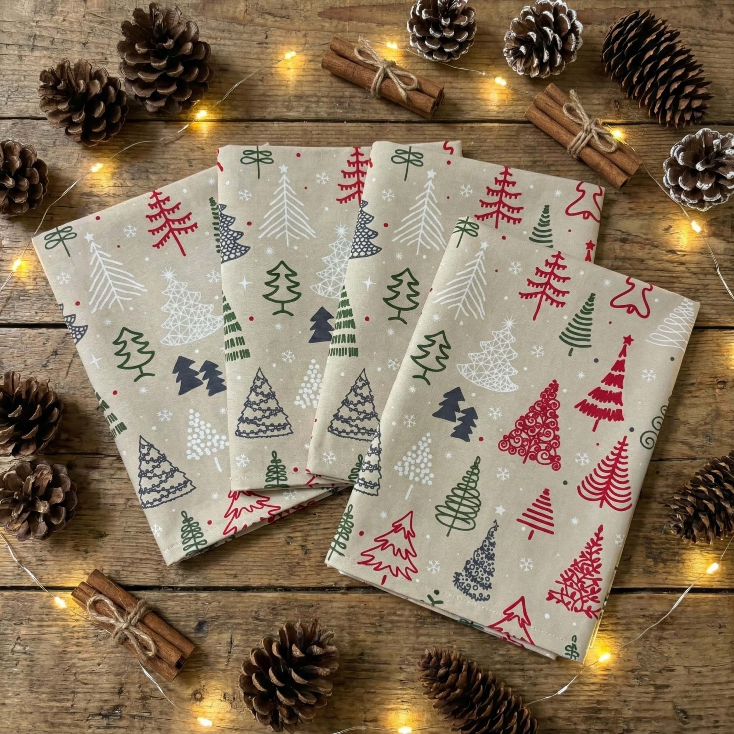 Four folded holiday napkins with Christmas tree patterns, surrounded by pinecones, cinnamon sticks, and fairy lights on a wooden surface.