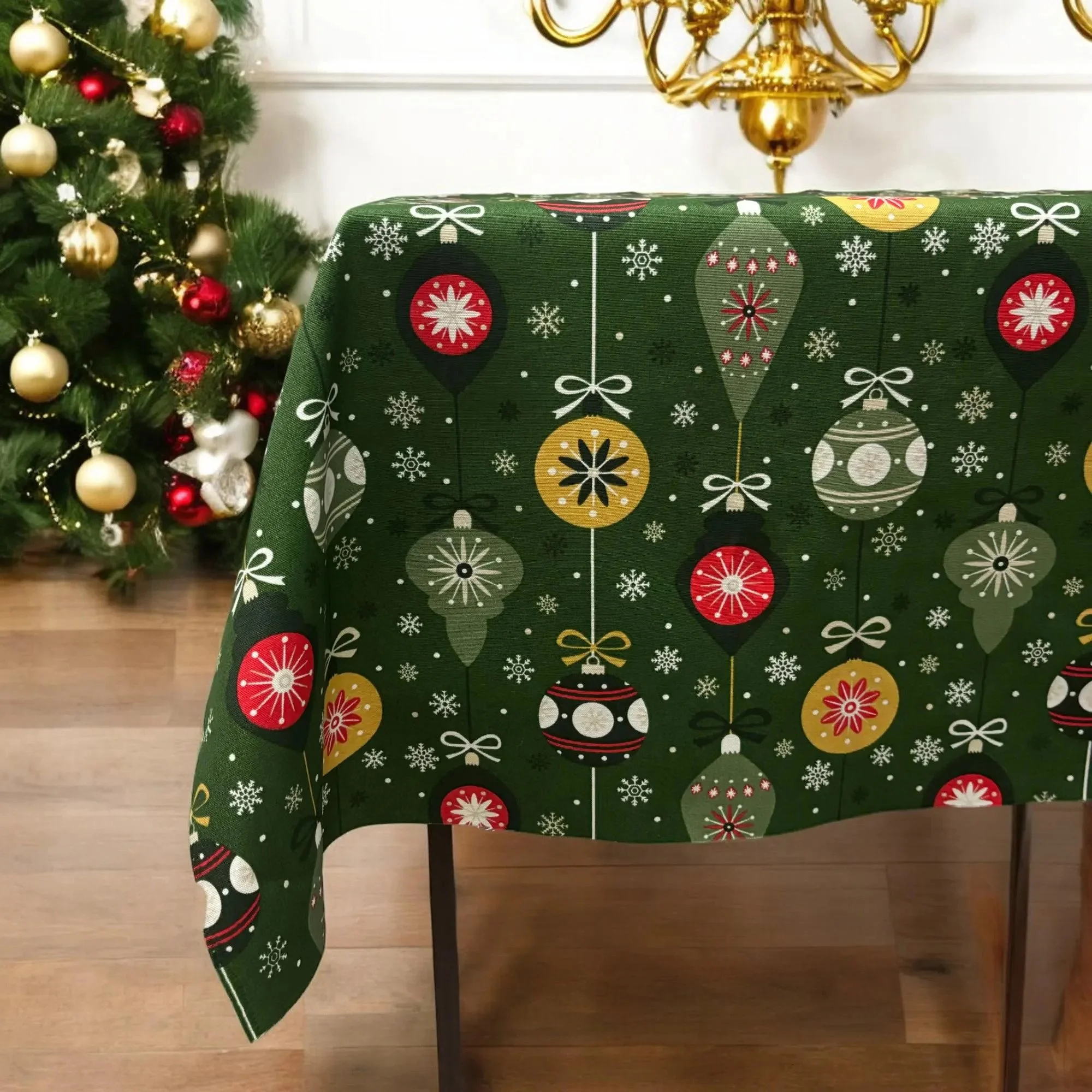 A Christmas-themed tablecloth with red, white, green, and gold ornaments on a dark green background, draped over a table, with a decorated Christmas tree with gold and red baubles in the background.