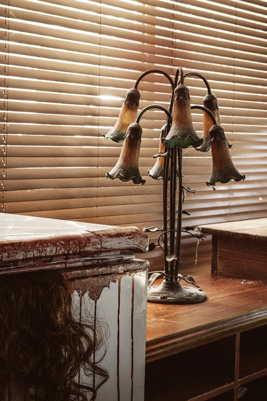 A vintage floor lamp with multiple flower-shaped lampshades, and a piece of furniture with a wet, dark red stain and hair strands on its surface, set against closed horizontal blinds.