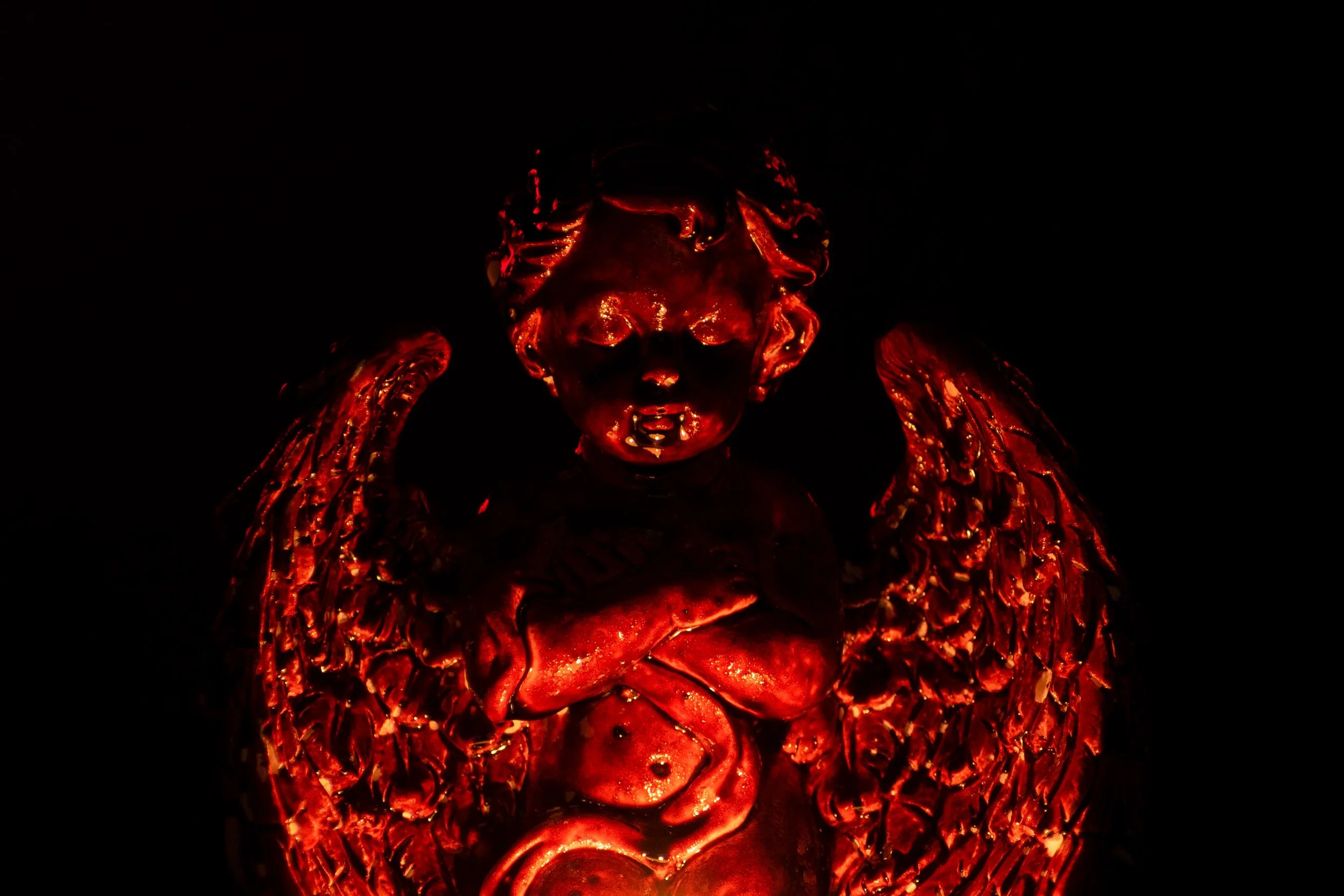 A glowing red sculpture of a cherub with wings, crossed arms, and a serpent coiled around its waist, illuminated against a dark background.
