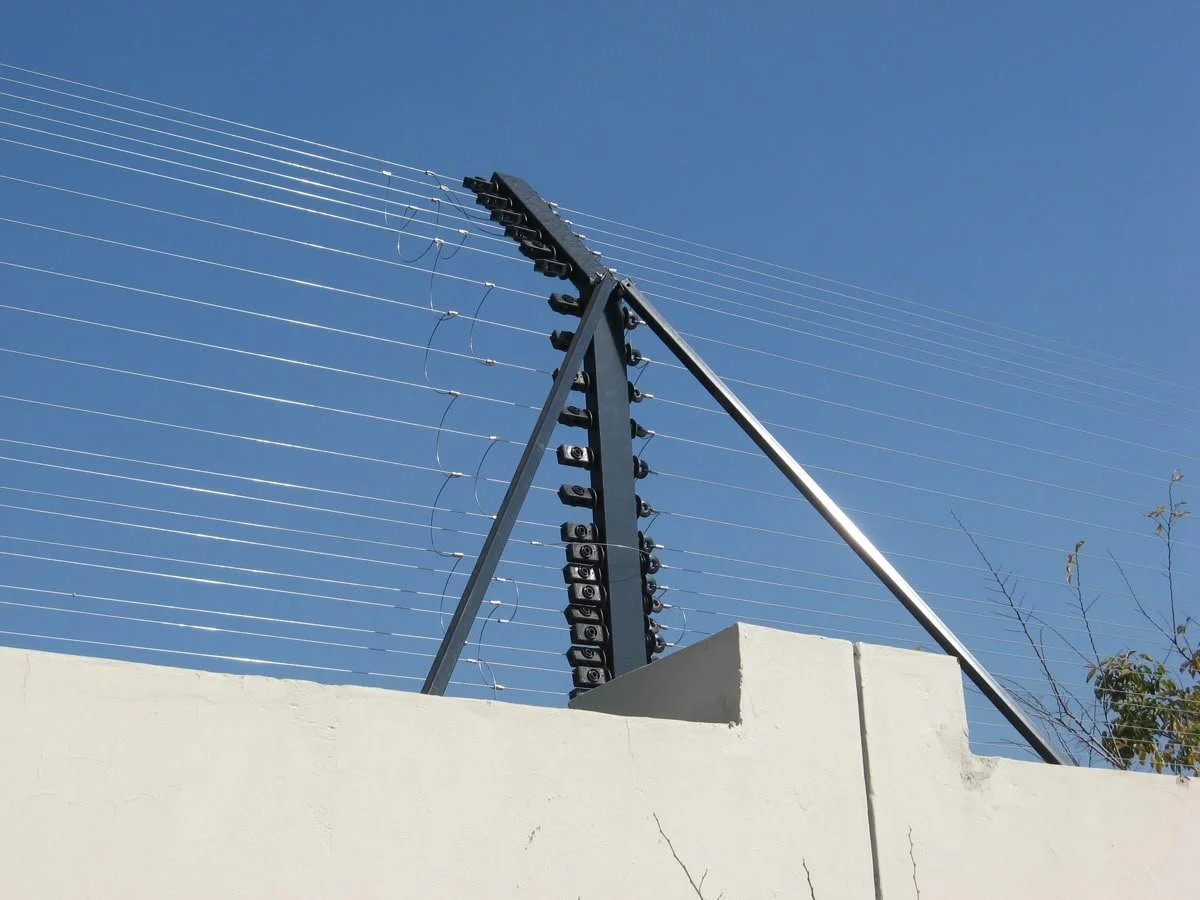 Security camera and barbed wire on top of a white wall against a clear blue sky.