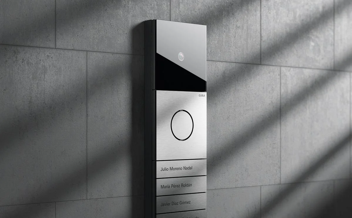 A modern intercom system mounted on a gray tiled wall with a circular button and a list of three names below it.
