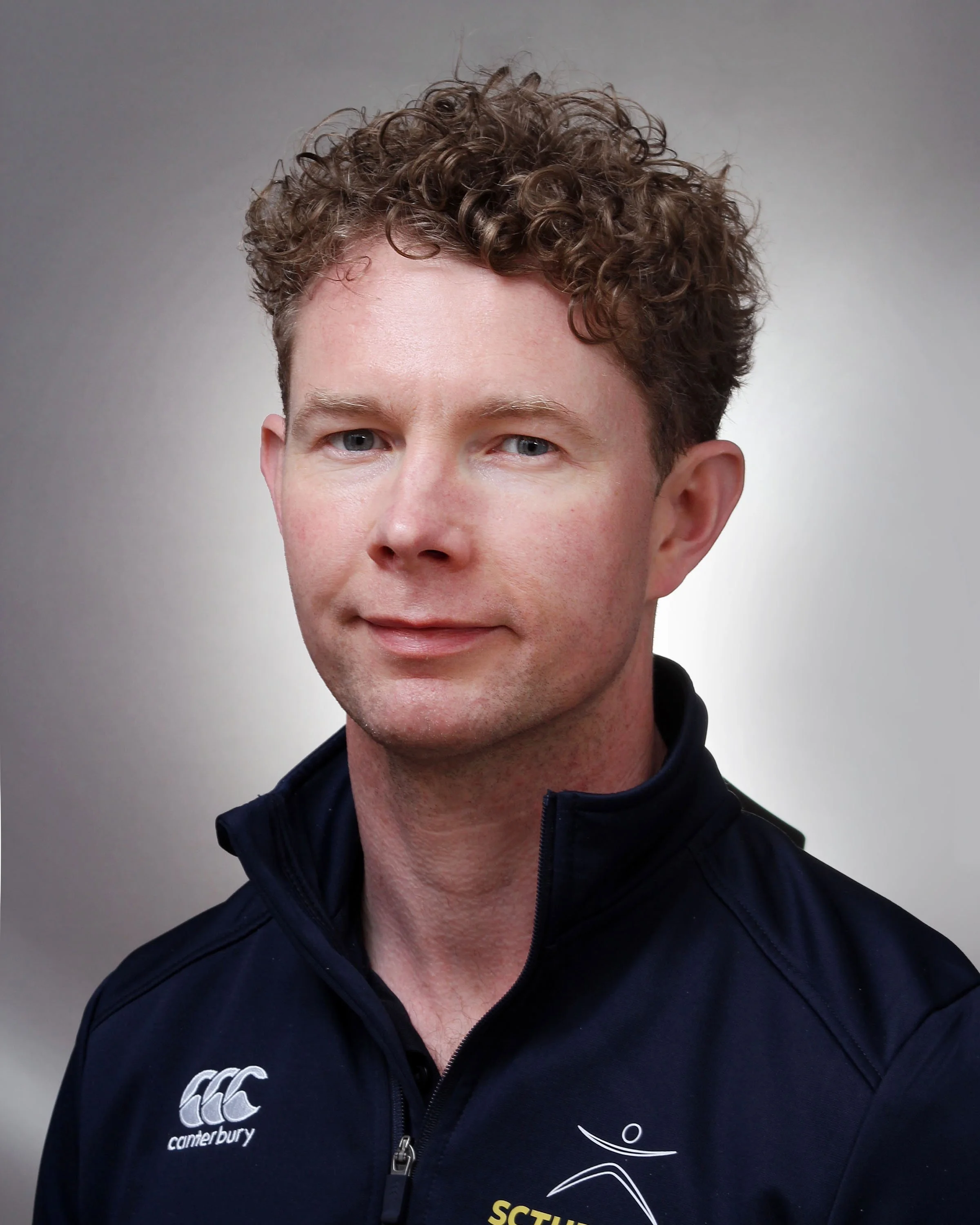 Portrait of a man with curly brown hair wearing a dark blue sports jacket with the Canterbury logo and SCTE logo.
