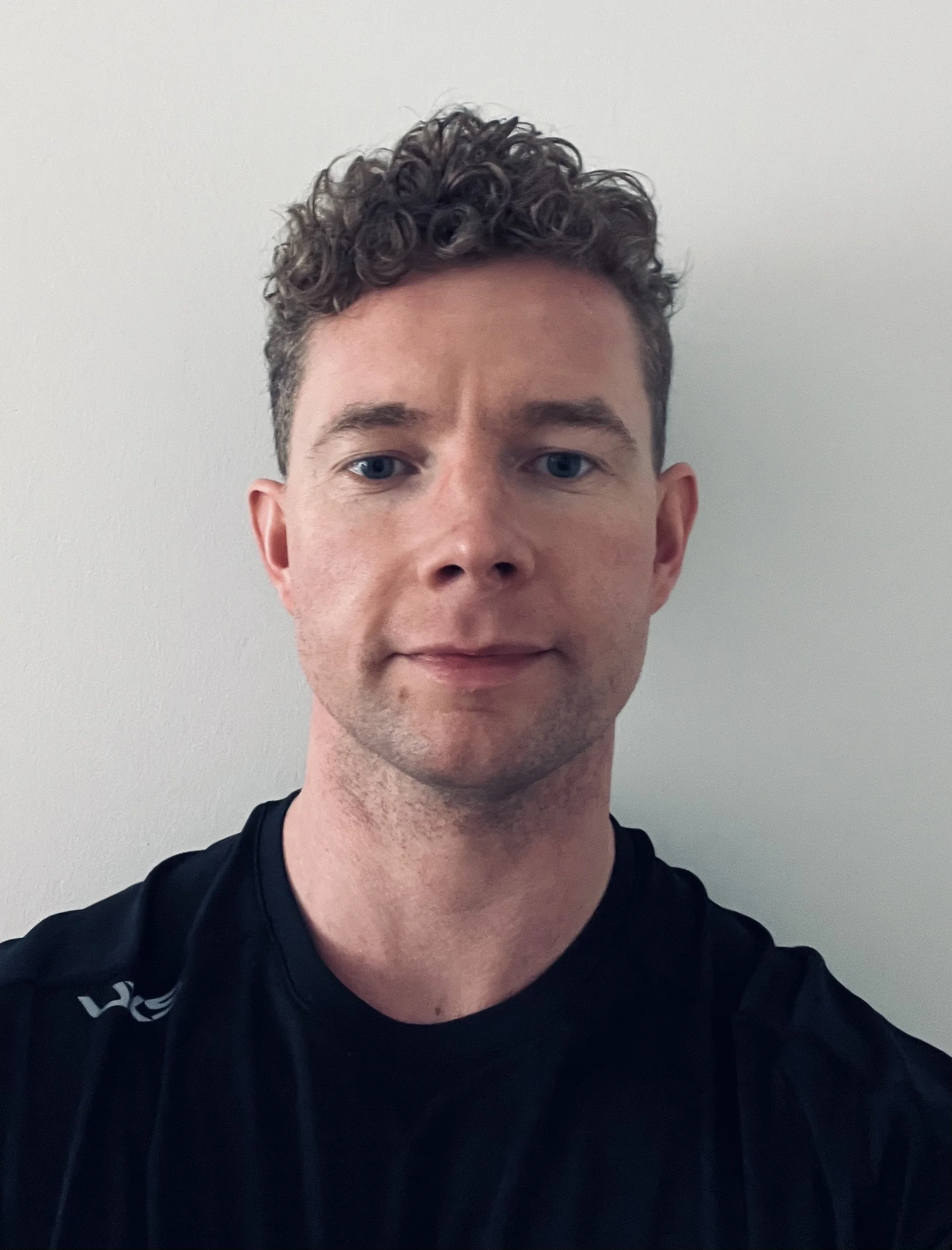 A young man with curly brown hair and blue eyes wearing a black t-shirt standing against a plain white wall.