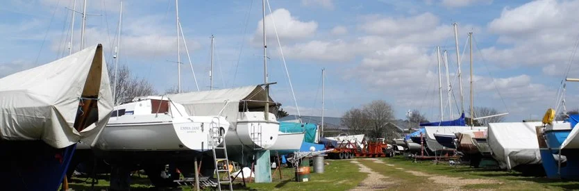 Sailboats on land at a marina, with some covered and others uncovered, under a partly cloudy sky.