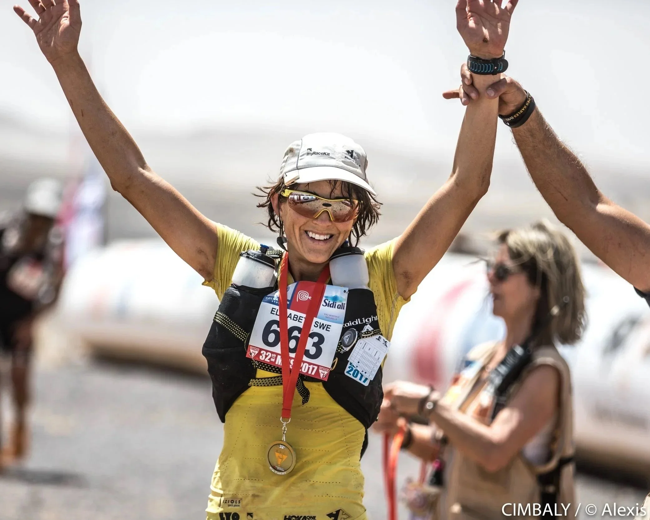 Elisabet Barnes celebrating her victory in the Marathon des Sables, wearing a yellow shirt, sunglasses, a cap, with a medal around her neck, holding her arms up in triumph, and being congratulated by someone off-camera.