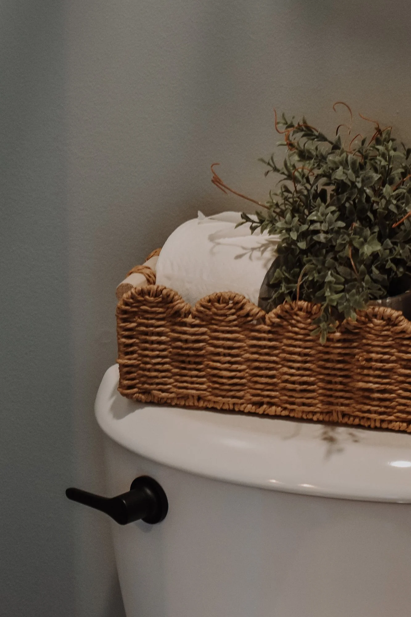 rustic modern farmhouse bathroom details,  wicker backet, greenery toilet paper storage above simple toliet with black insdustrial handle