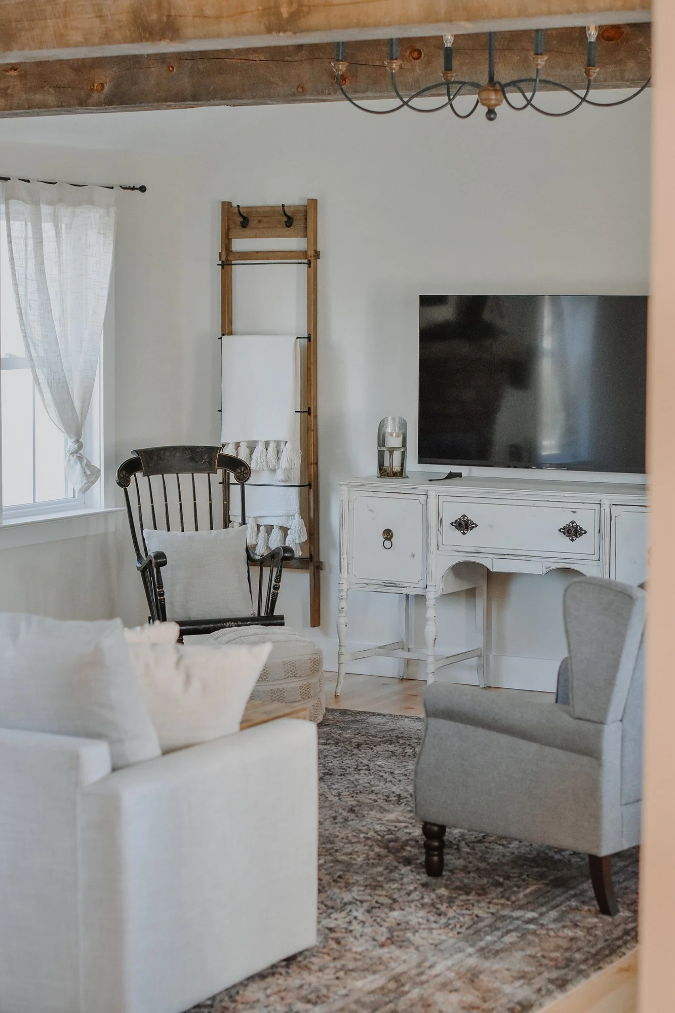 1890 restored farmhouse entry room high ceiling, antique candlestick chandelier, reclaimed wood beams, farmhouse decor, neautral cozy sitting area with couch and arm chairs, rocking chair, blanket ladder, antiquue buffet anf flat screen tv