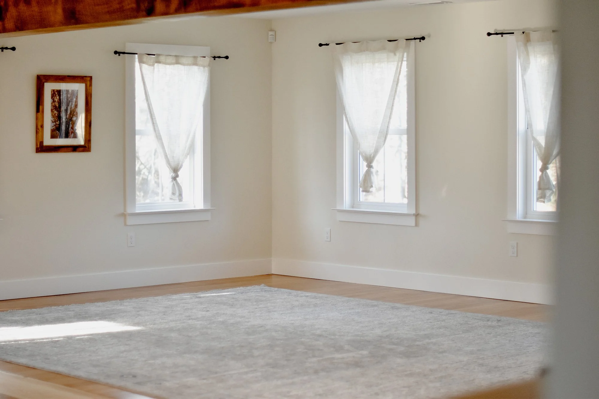 1890 restored farmhouse dining room small gathering space lined with windows pouring in natural light, reclaimed wood beams and refinished original wood floors, creamy opaque tied linen curtains line the wall, rustic rug, open space