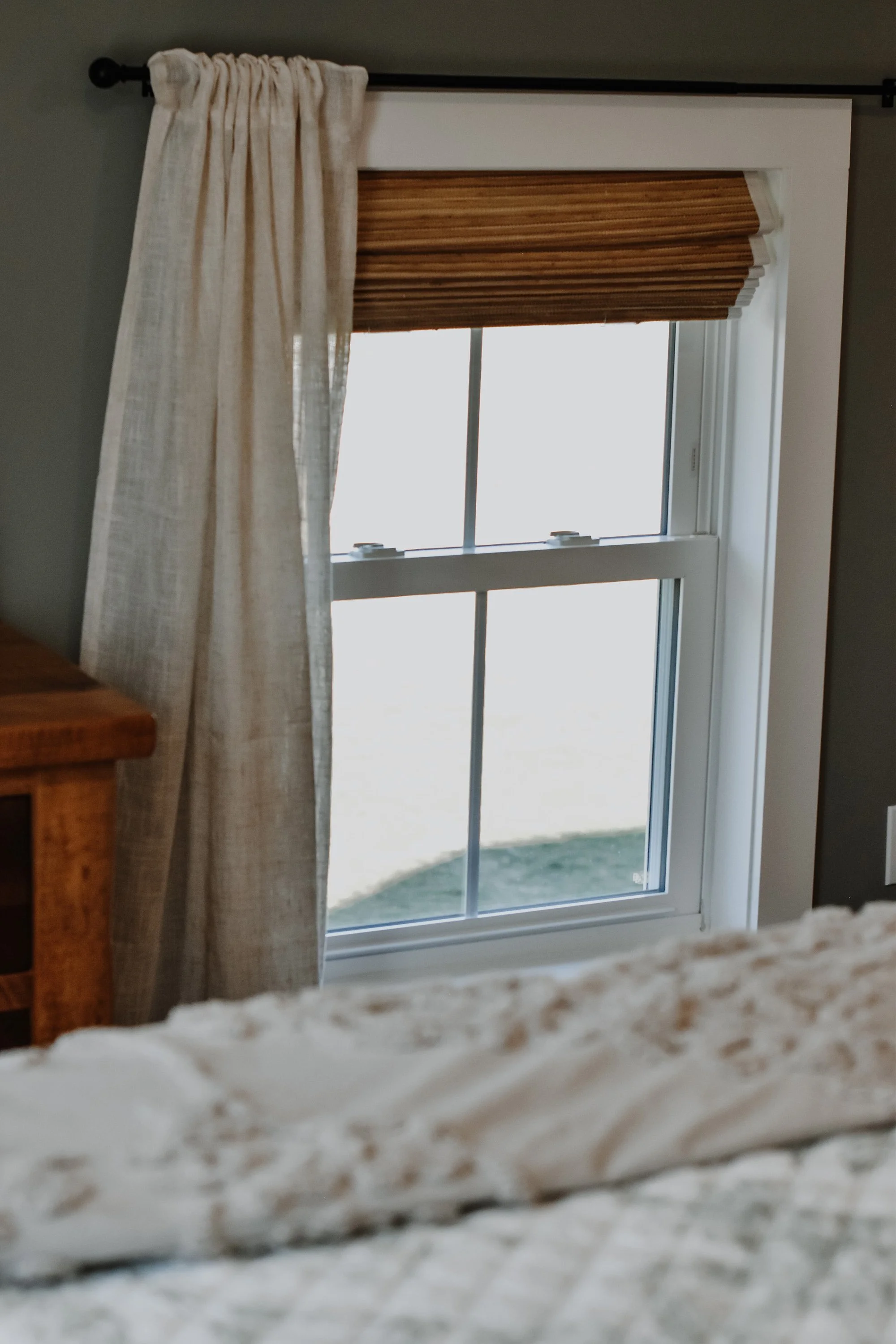 1890 restored farmhouse stay rustic master bedroom with solid wooden handcrafted bedframe, creamy floral bed linens, window with linen curtains, refurbished original farmhouse wood floors, antique wooden furtinute with lamp and framed art
