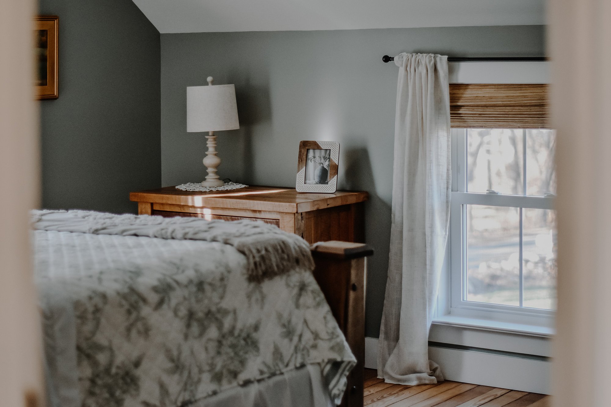 1890 restored farmhouse stay rustic master bedroom with solid wooden handcrafted bedframe, creamy floral bed linens, window with linen curtains, refurbished original farmhouse wood floors, antique wooden furtinute with lamp and framed art