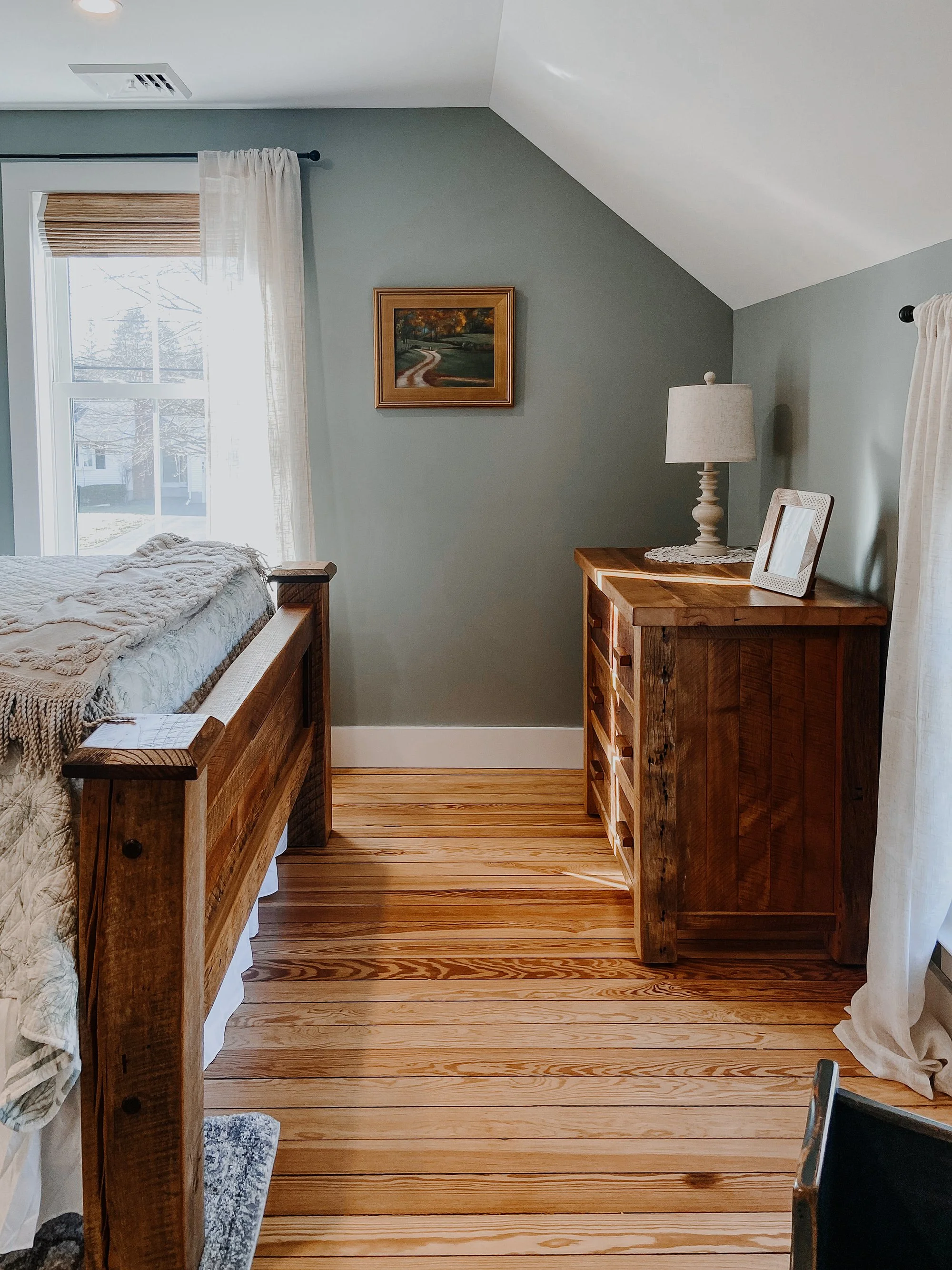 1890 restored farmhouse stay rustic master bedroom with solid wooden handcrafted bedframe, creamy floral bed linens, window with linen curtains, refurbished original farmhouse wood floors, antique wooden furtinure with lamp and framed art