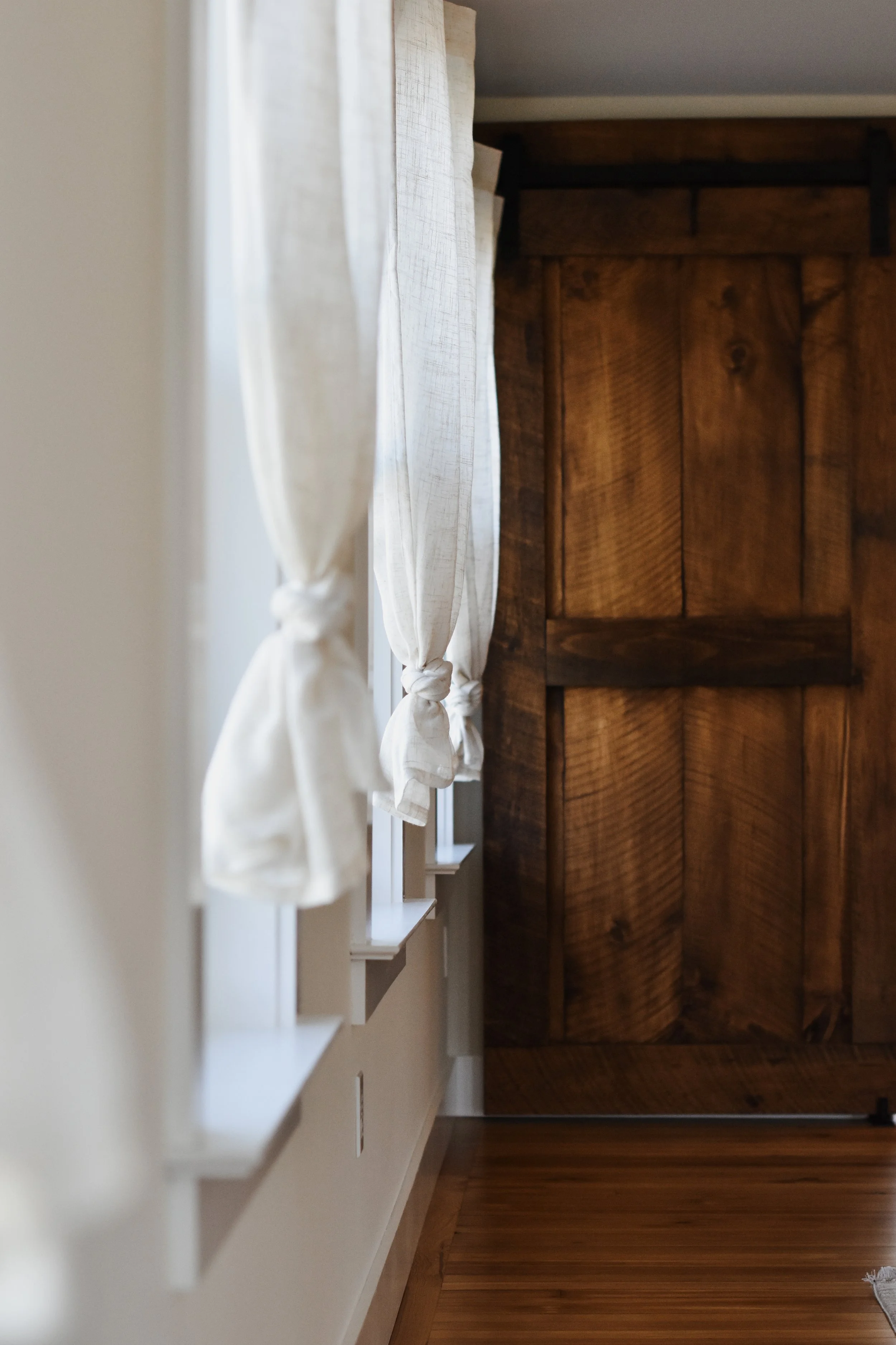1890 restored farmhouse dining room small gathering space lined with windows pouring in natural light, reclaimed wood farm doors and refinished original wood floors, creamy opaque tied linen curtains line the wall