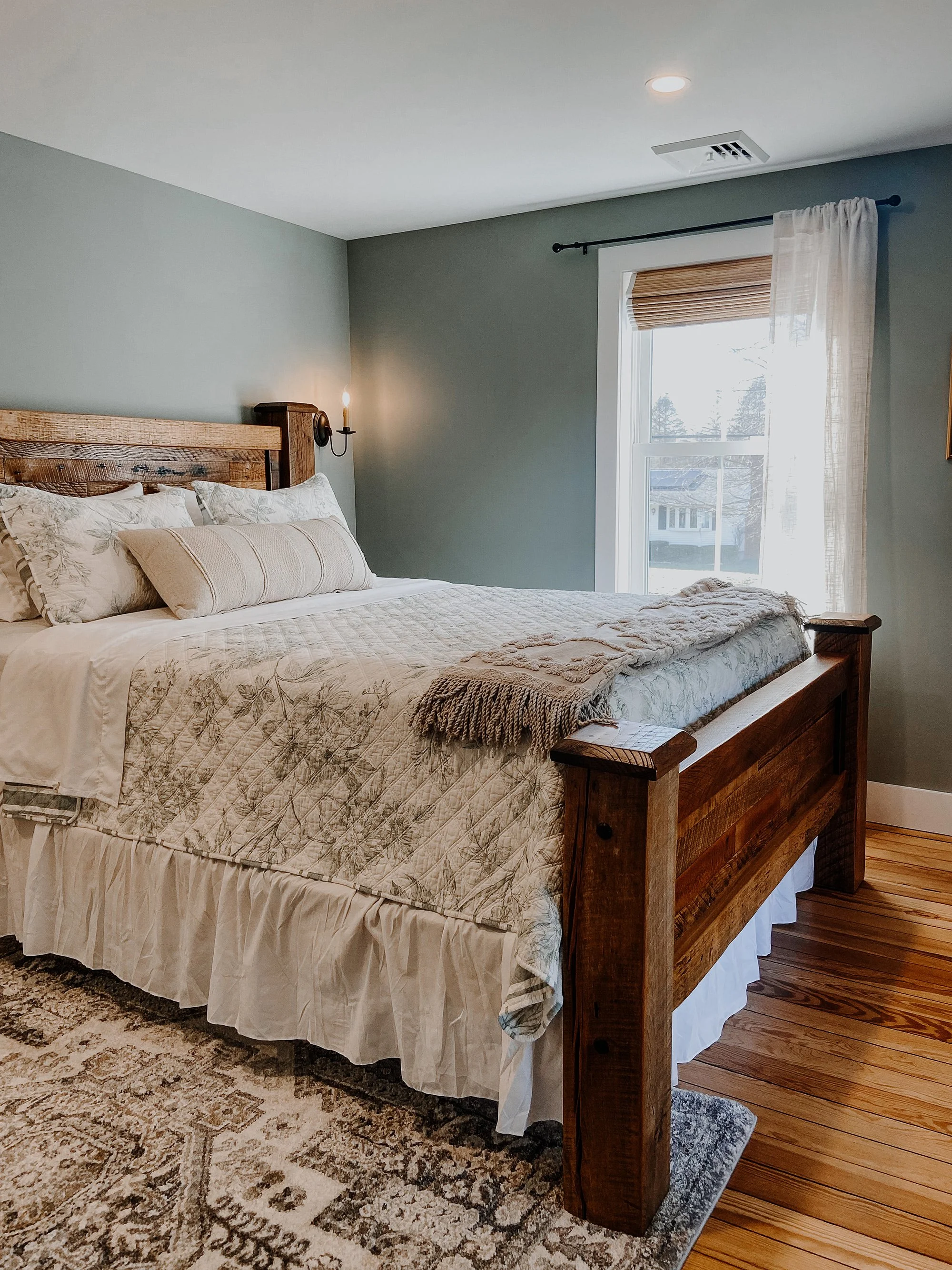 1890 restored farmhouse stay rustic master bedroom with solid wooden handcrafted bedframe, creamy floral bed linens, candlestick light fixtures, window with linen curtains, refurbished original farmhouse wood floors, rustic rug