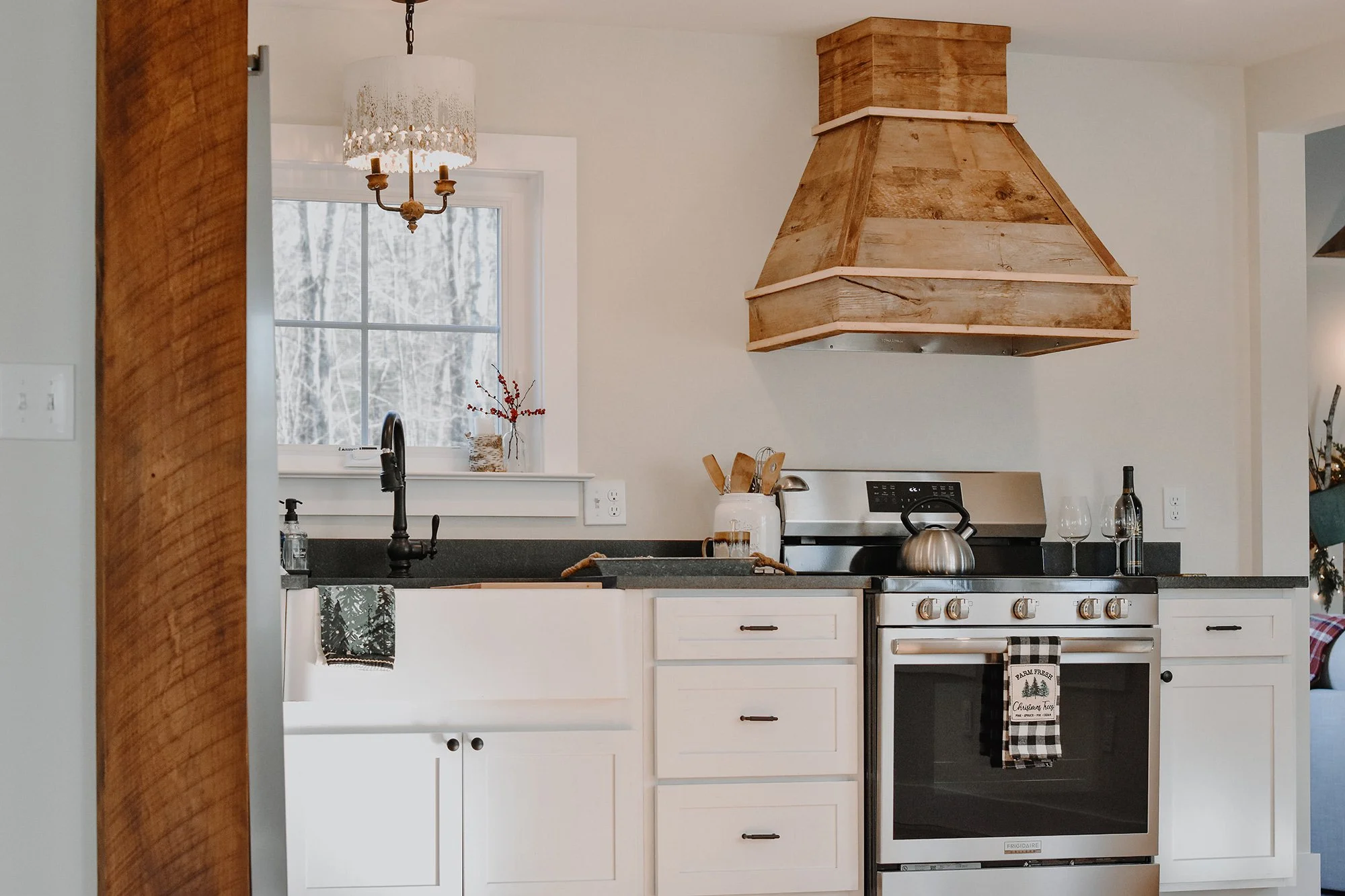 1890 restored farmhouse stay kitchen with modern and rustic details, creamy white, new cabinets, antique chandelier, reclaimed wood oven hood, above the sink window,  modern stainless steel oven, tea kettle, wine, kitchen utensils