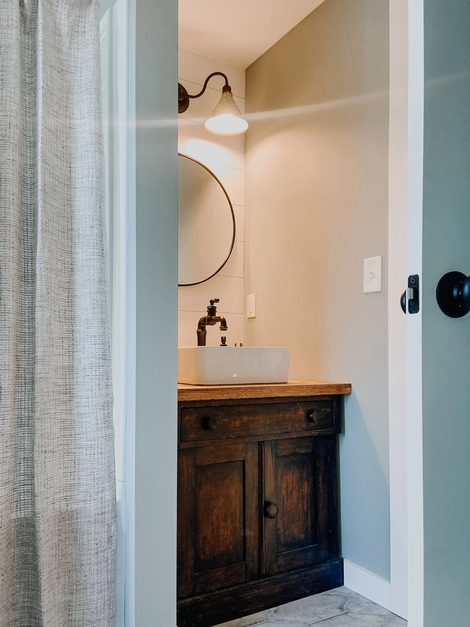 rustic modern farmhouse bathroom tile floor, shower with modern fixtures and cotton shower curtain, refurbished undersink cabinet, farmhouse sink, industrial sink fixture, circle mirror, metal cone sconces lighting