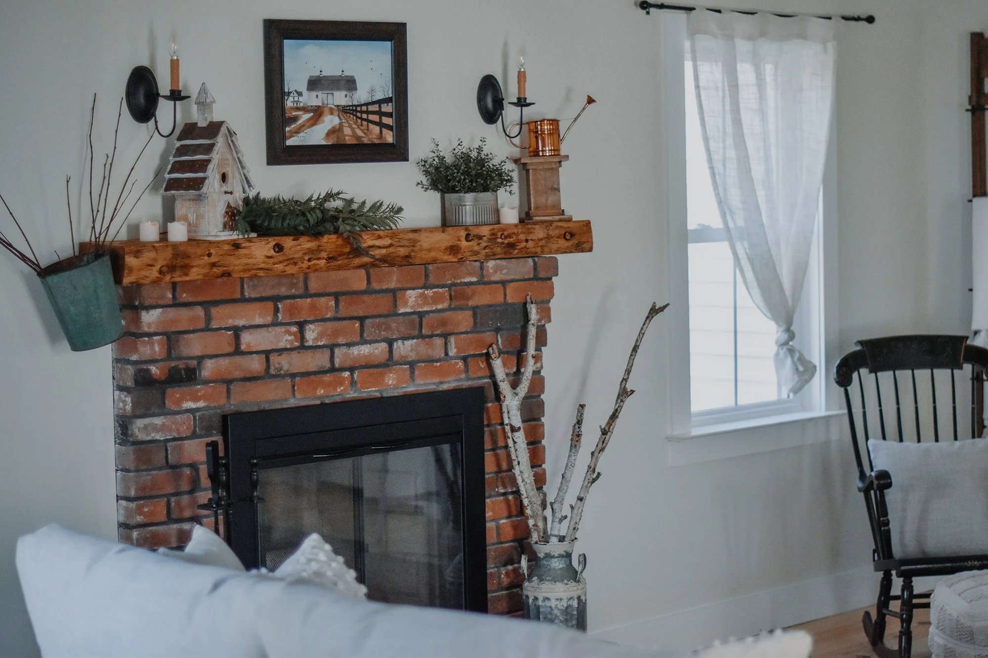 1890 restored farmhouse stay gathering room original brick wood burning fireplace, reclaimed wood mantal, birch branck decor, birhouse, copper watering can, antique rocking chair, candlestick light fixture, and cozy sitting