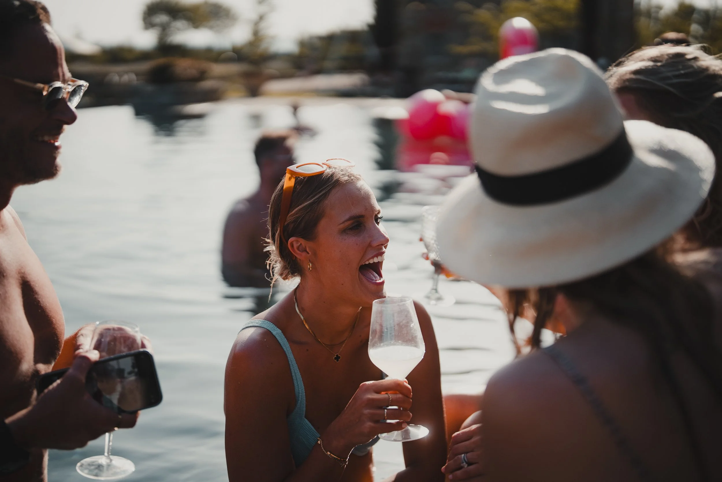 A group of people enjoying a social gathering near a body of water, holding glasses of wine, with some wearing hats and sunglasses, during sunny weather.