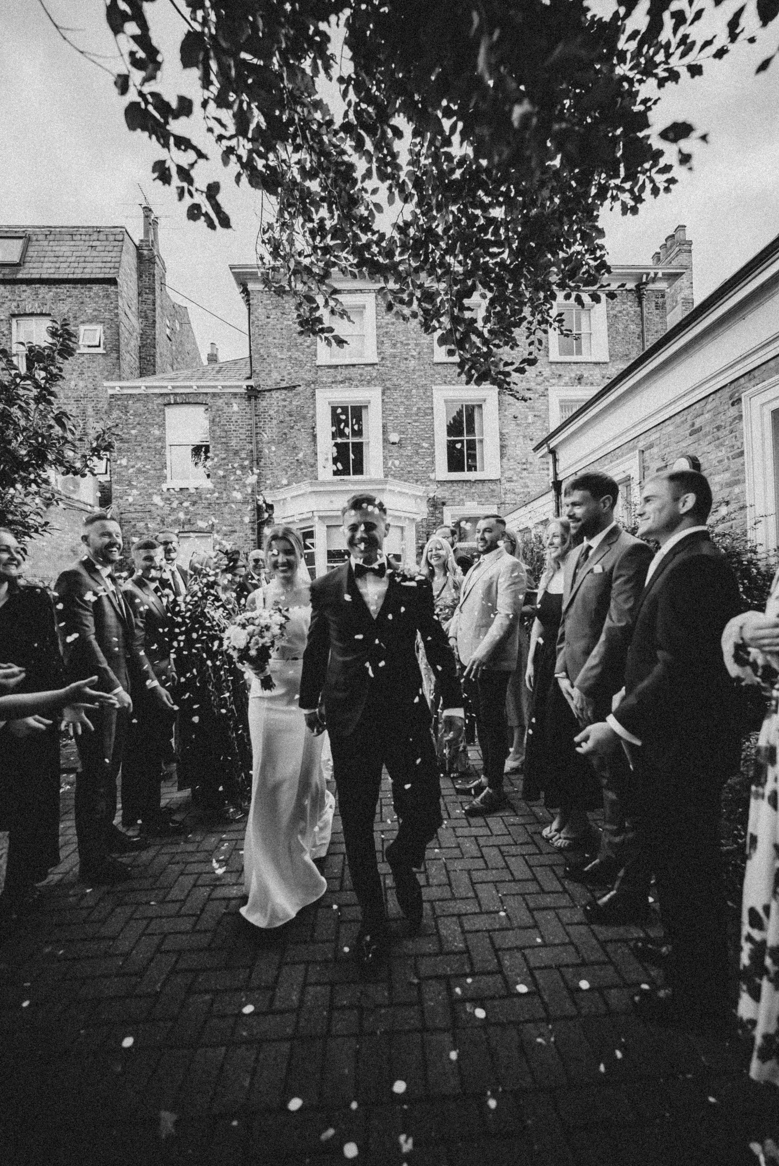 A black and white photo of a wedding celebration with a bride and groom walking hand in hand through a crowd of guests. The crowd is smiling and throwing confetti. There are multi-story brick buildings in the background and trees overhead.