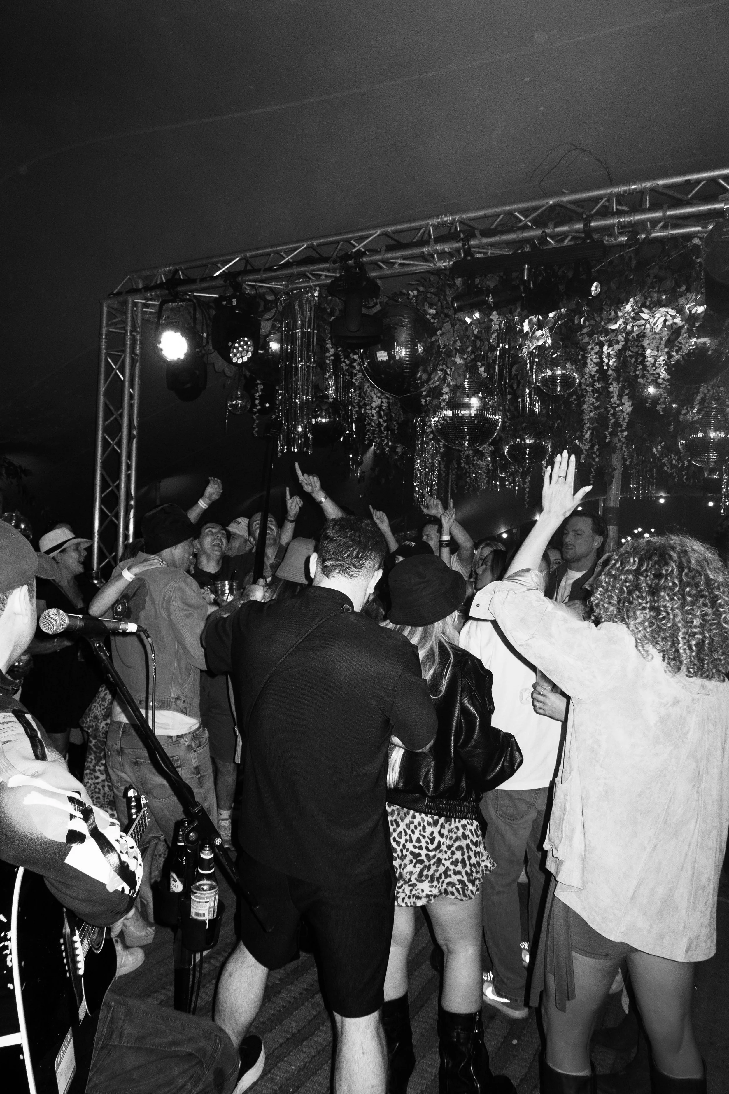 A lively indoor party with people dancing and enjoying music, decorated with disco balls, hanging ornaments, and lighting, with a stage and musical equipment.