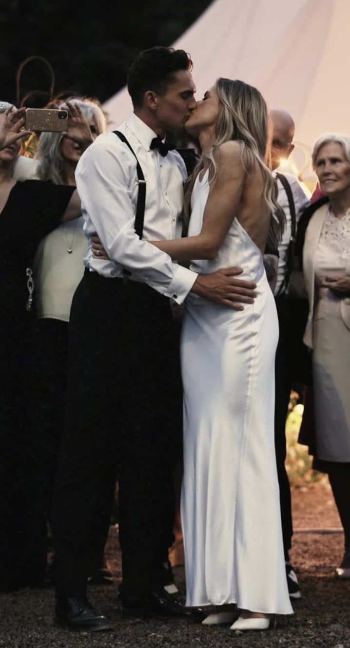 A couple in wedding attire kissing during their wedding ceremony, with guests watching and taking pictures in the background.
