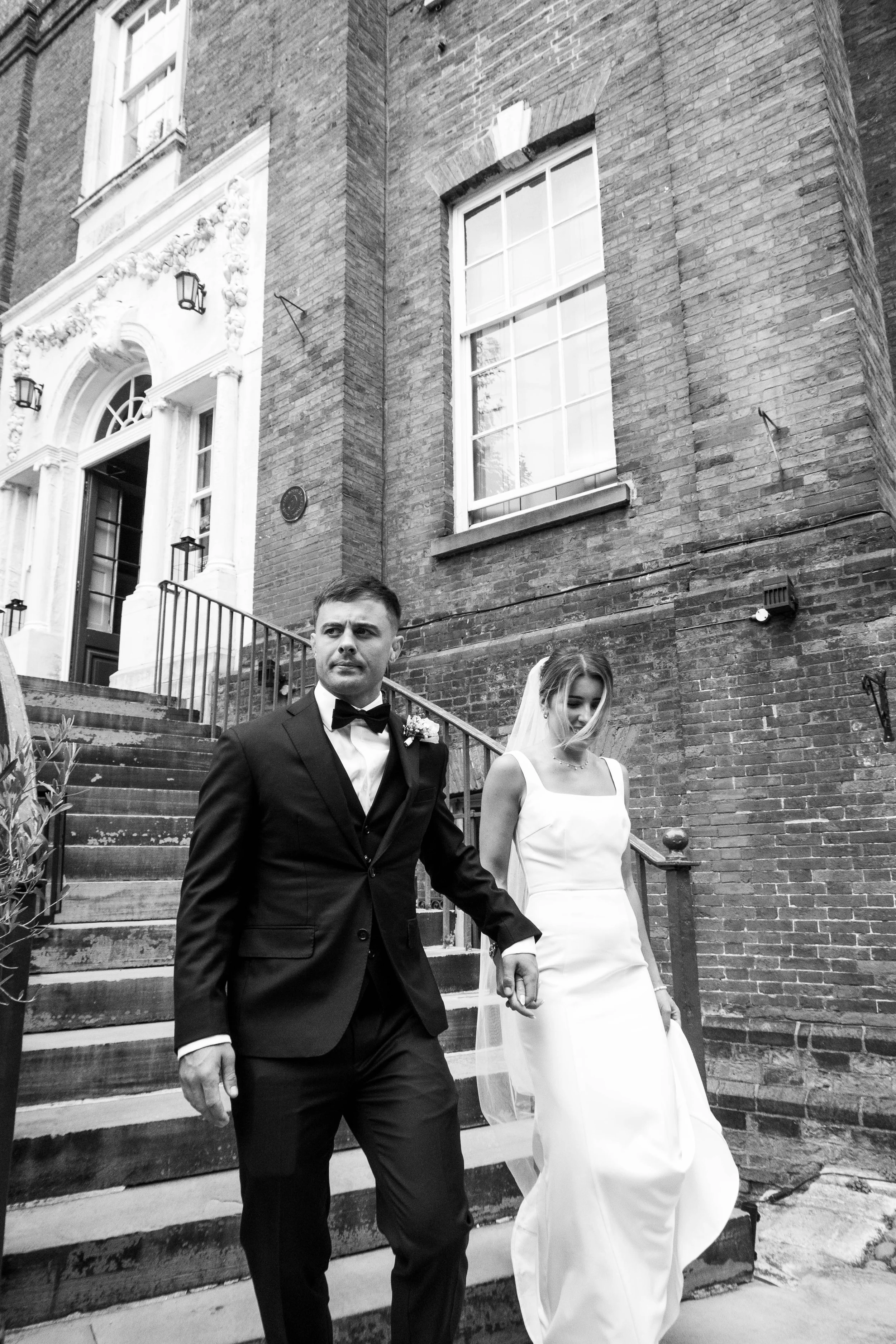 A bride and groom walking down the stairs in front of a brick building with large windows and decorative moldings.