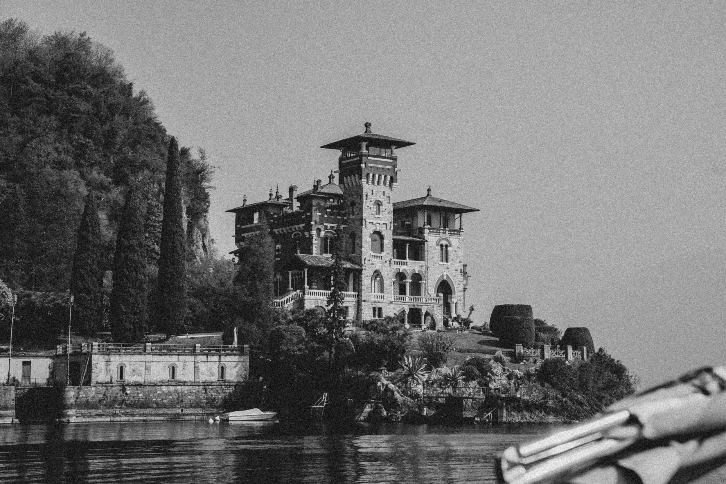 A black and white photo of a large castle-like house on a hill beside a body of water, with trees on the hill and small boats on the water.