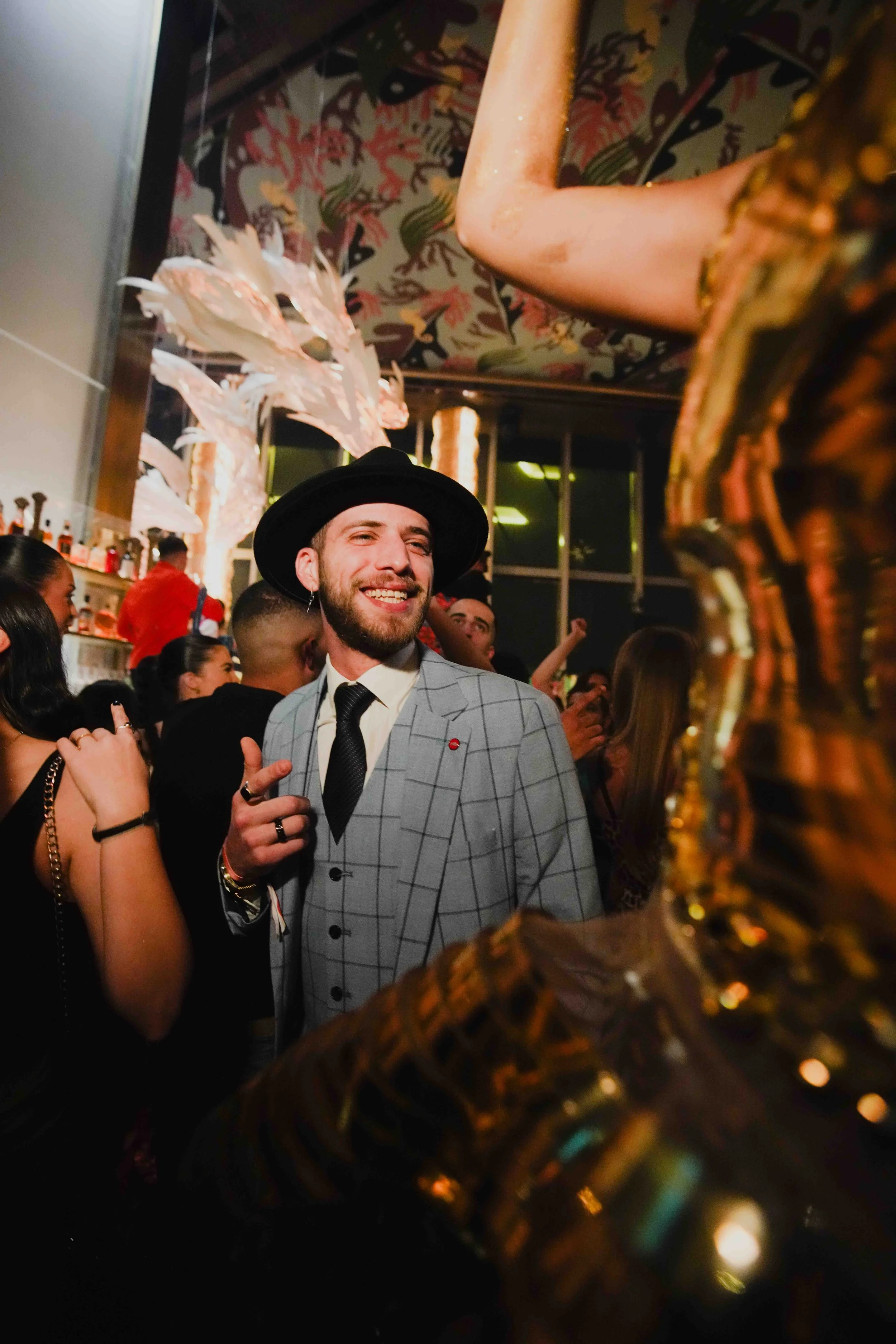 A smiling man wearing a black hat, gray checkered suit, and black tie at a lively indoor party with people dancing and a bar in the background. The photo is taken through a decorative golden object.